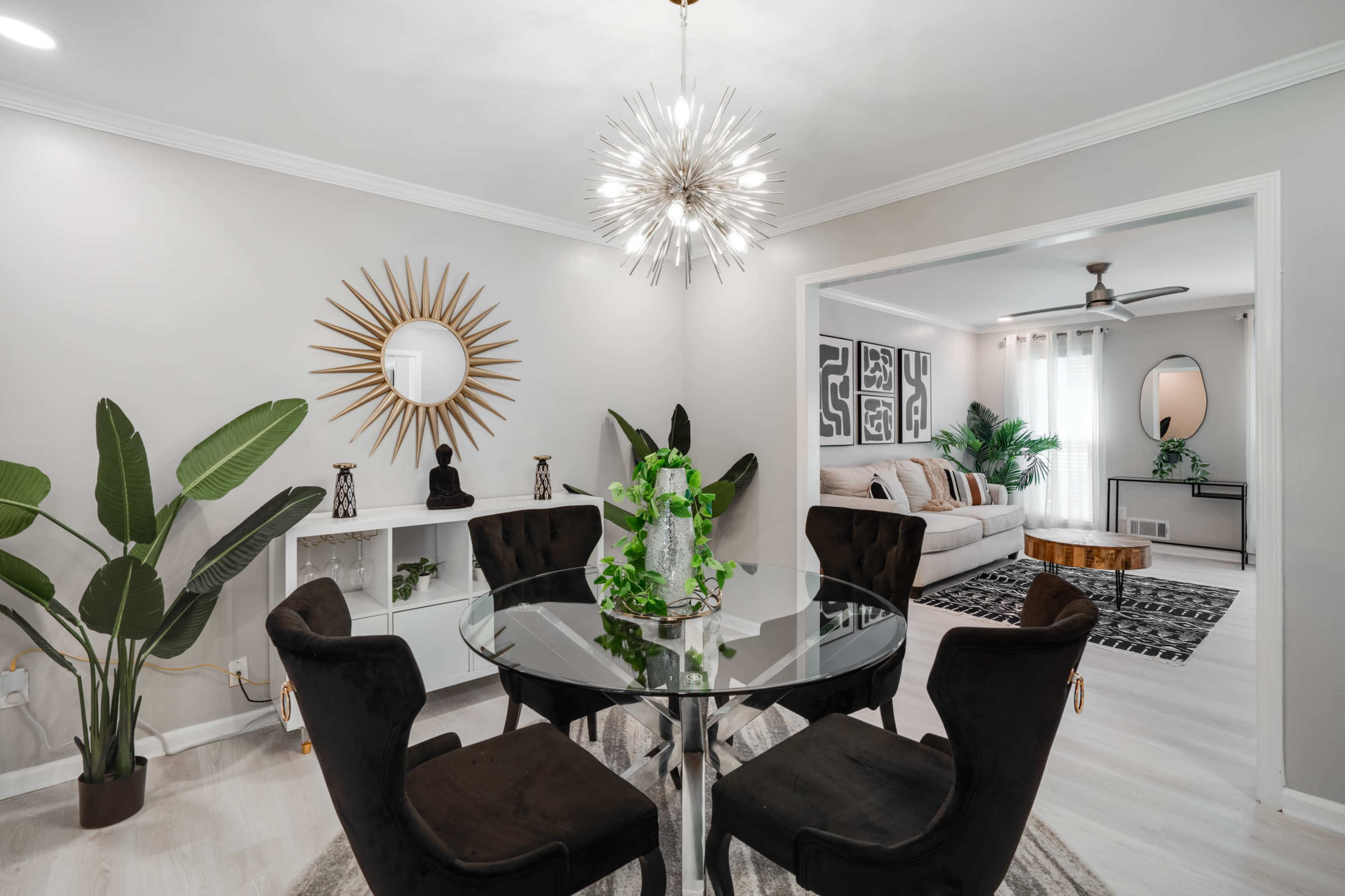 A modern dining area features a round glass table surrounded by black chairs, with decorative plants and a starburst chandelier above.