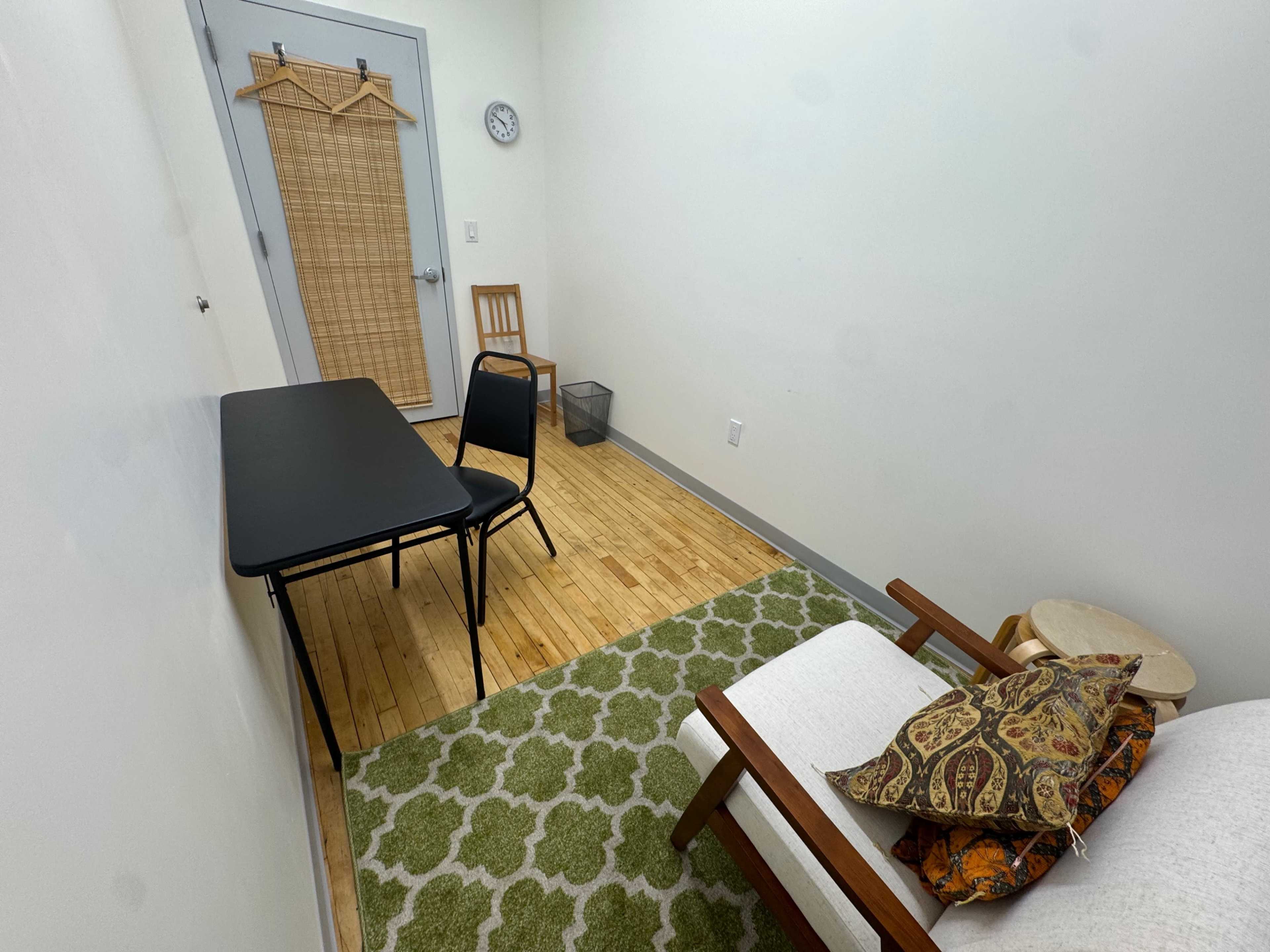 The image shows a small, sparsely furnished room with a table and chair, a wastebasket, and a couch with cushions on a patterned rug.