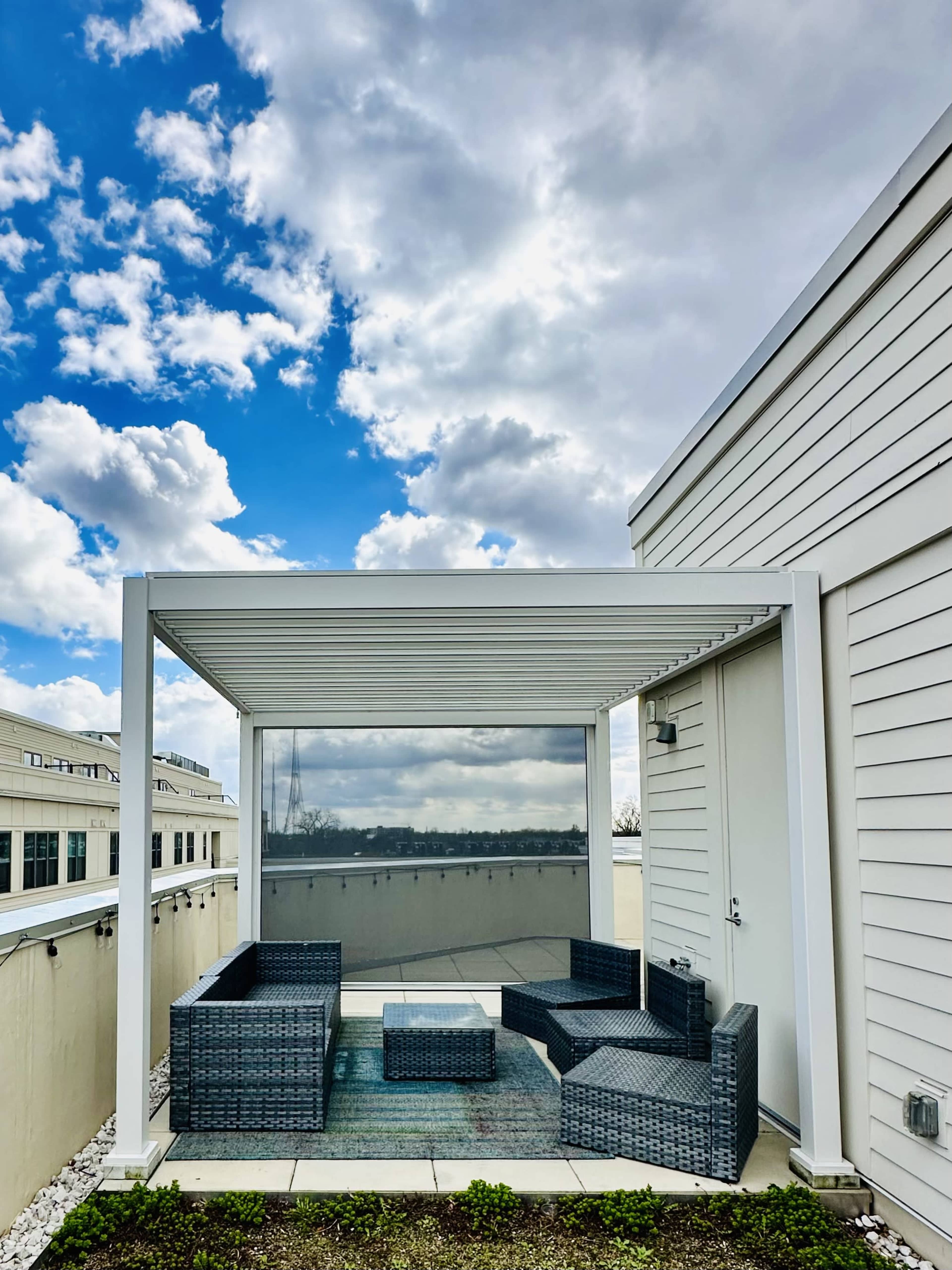 A covered rooftop patio features a set of gray wicker furniture and is surrounded by a bright sky with clouds.