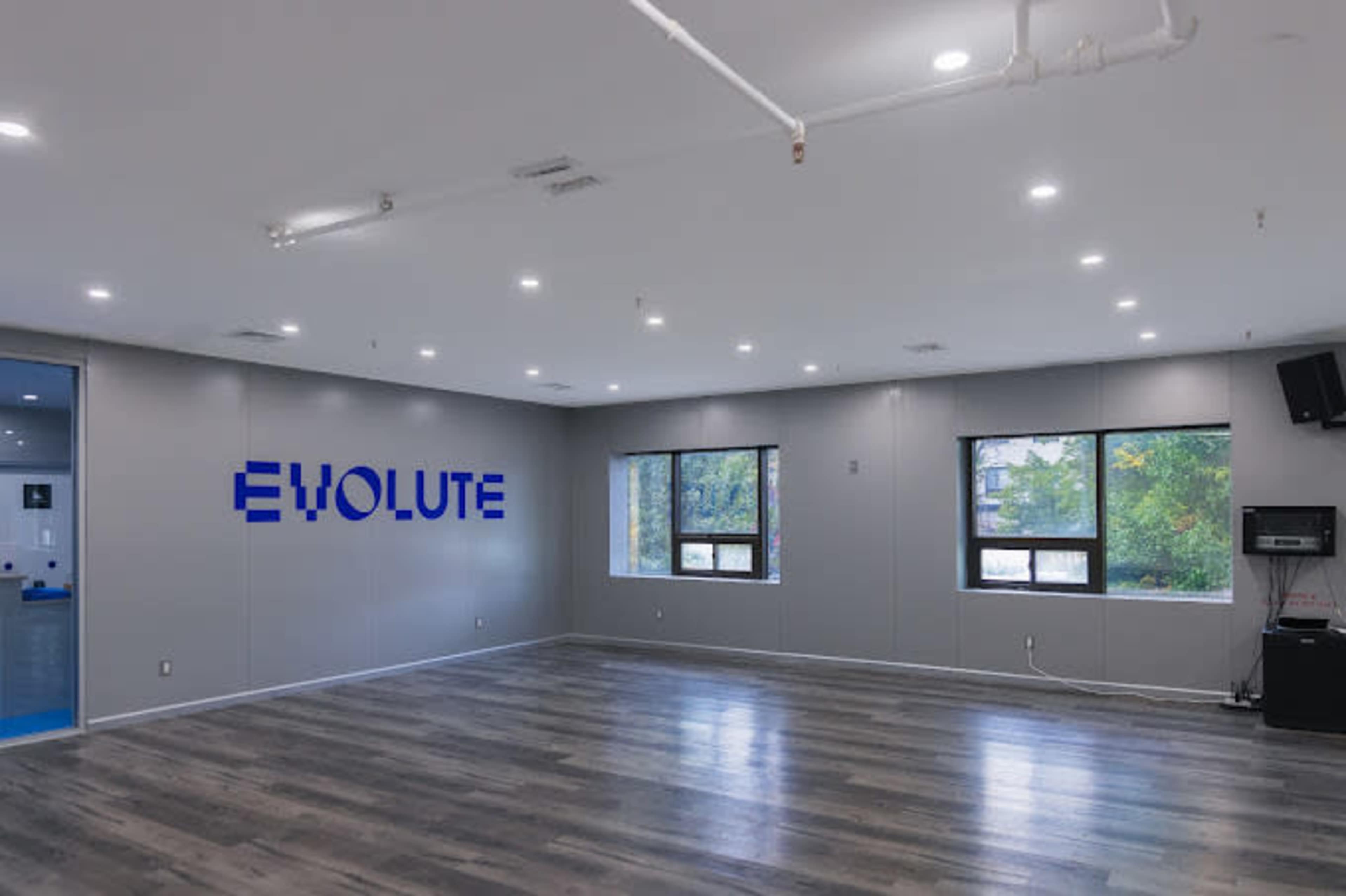 The image shows a spacious room with wooden flooring and large windows, featuring a blue "EVOLUTE" sign on the gray wall.