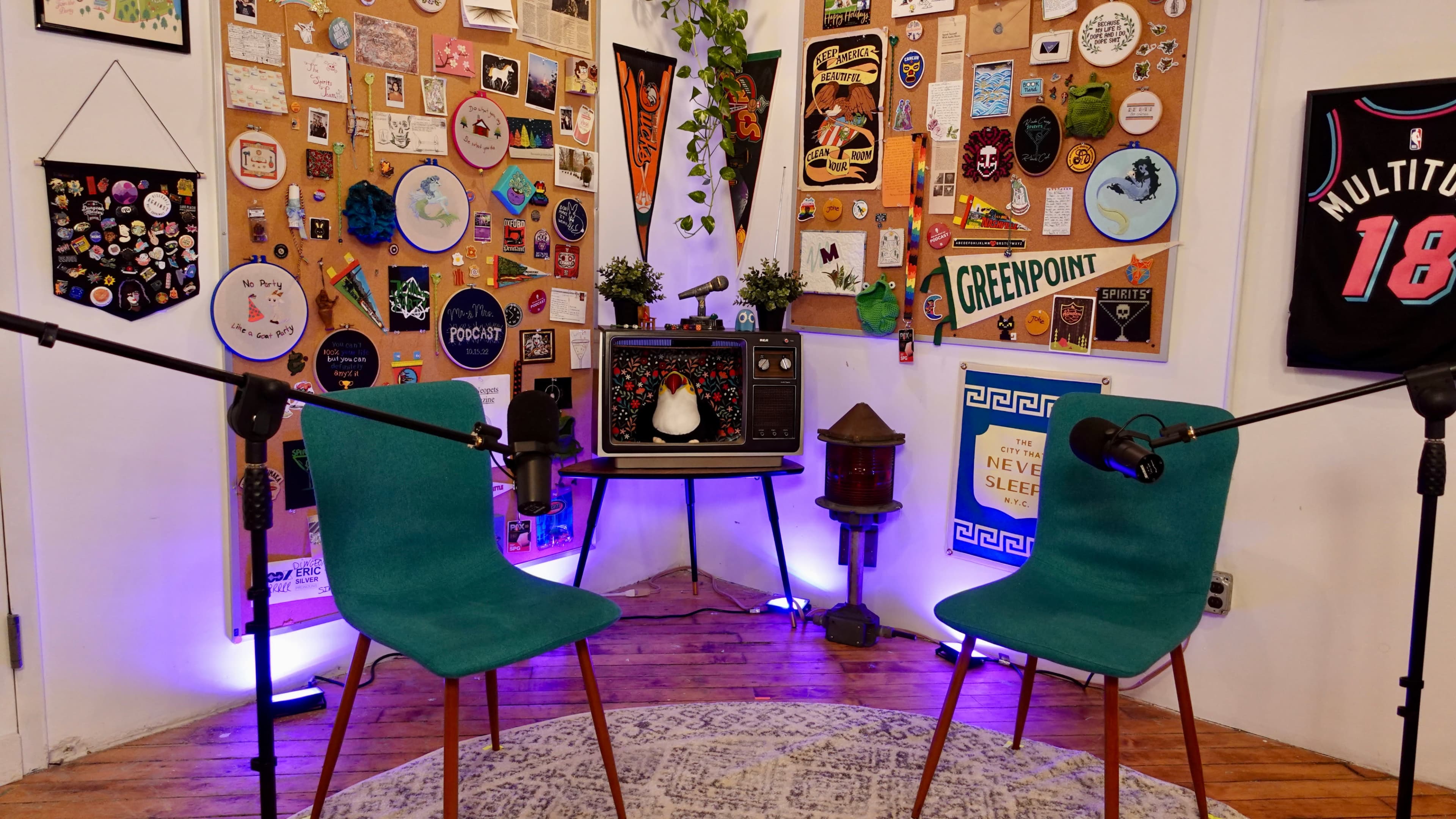 A cozy podcast or interview setup with two teal chairs facing a vintage-style display board adorned with various pins, posters, and decorations.