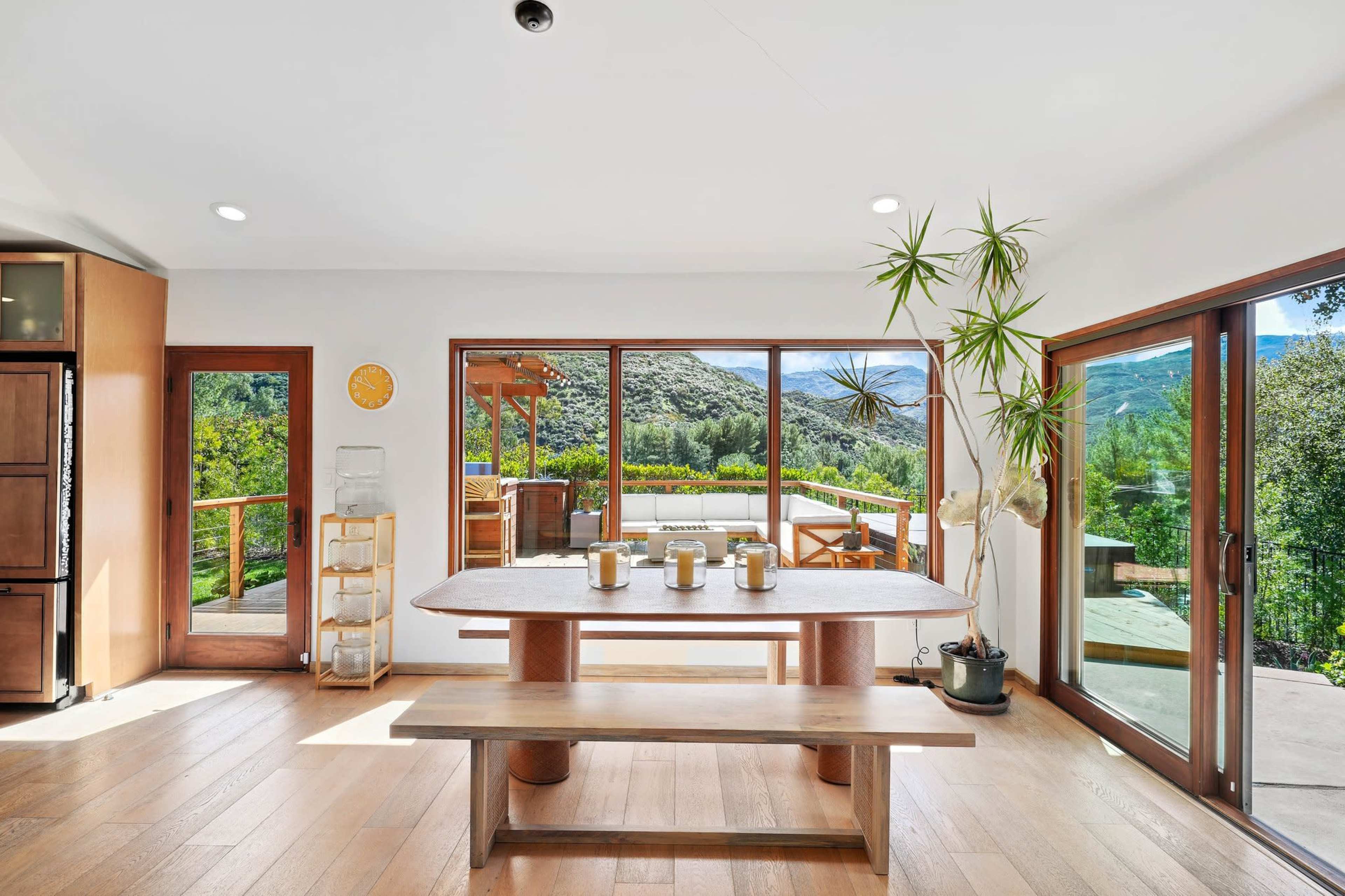 The image shows a modern dining area with a wooden table and a view of a green landscape through large sliding glass doors.