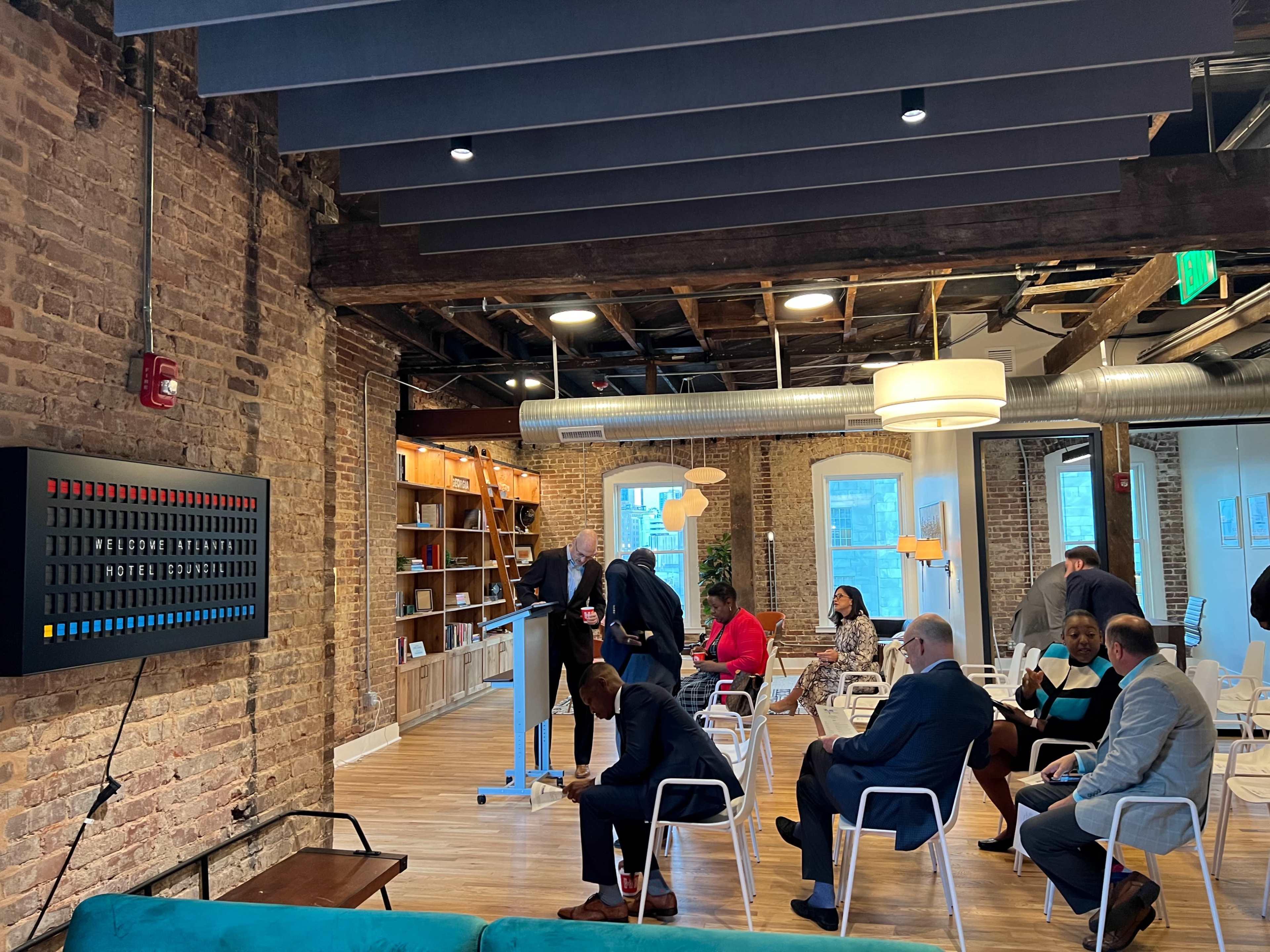 A group of people are gathered in a modern office space with exposed brick walls, some standing near a presentation setup while others are seated.