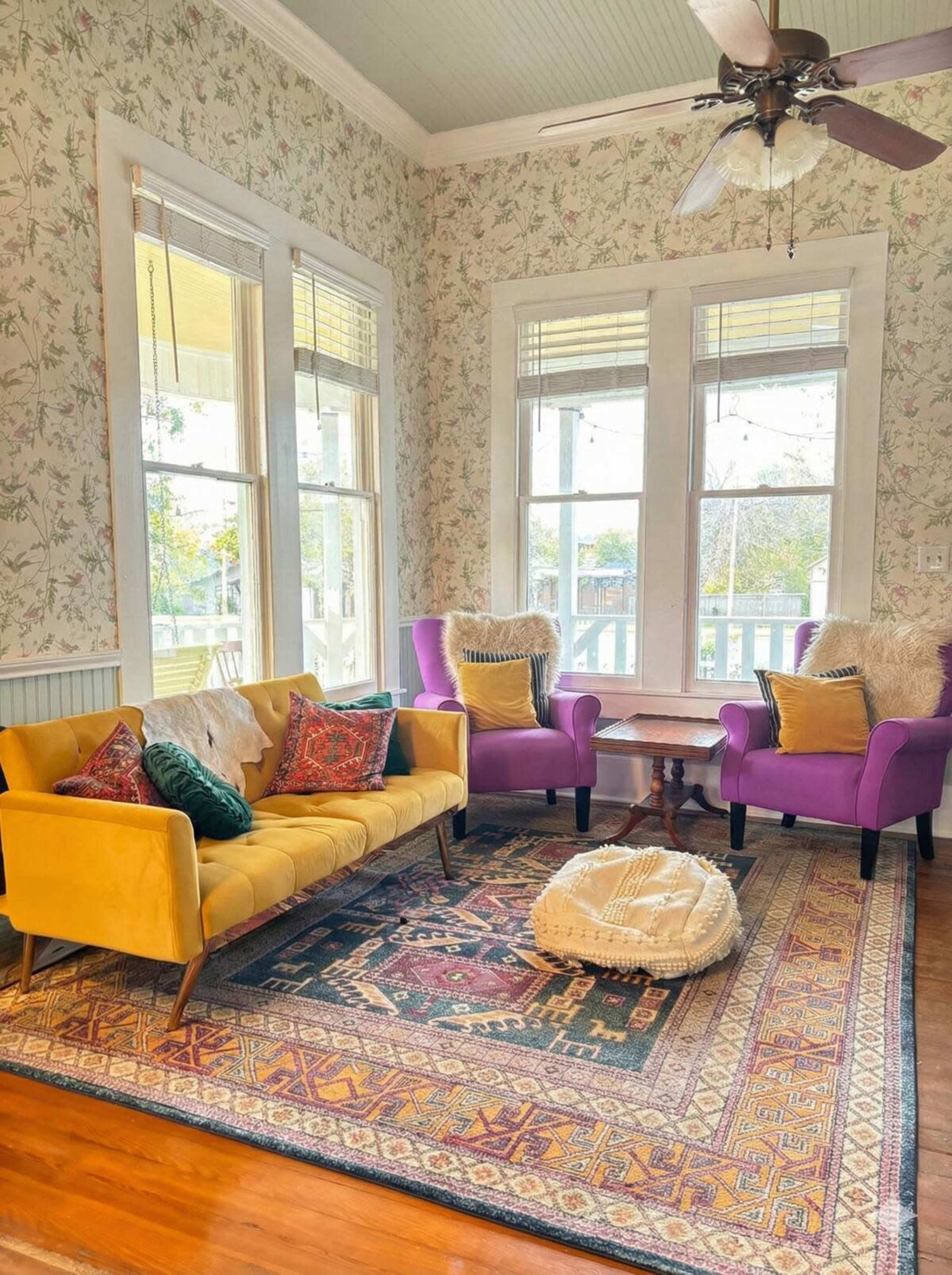 The image shows a brightly decorated living room with a yellow sofa, two purple armchairs, a wooden coffee table, and a patterned area rug on hardwood flooring.