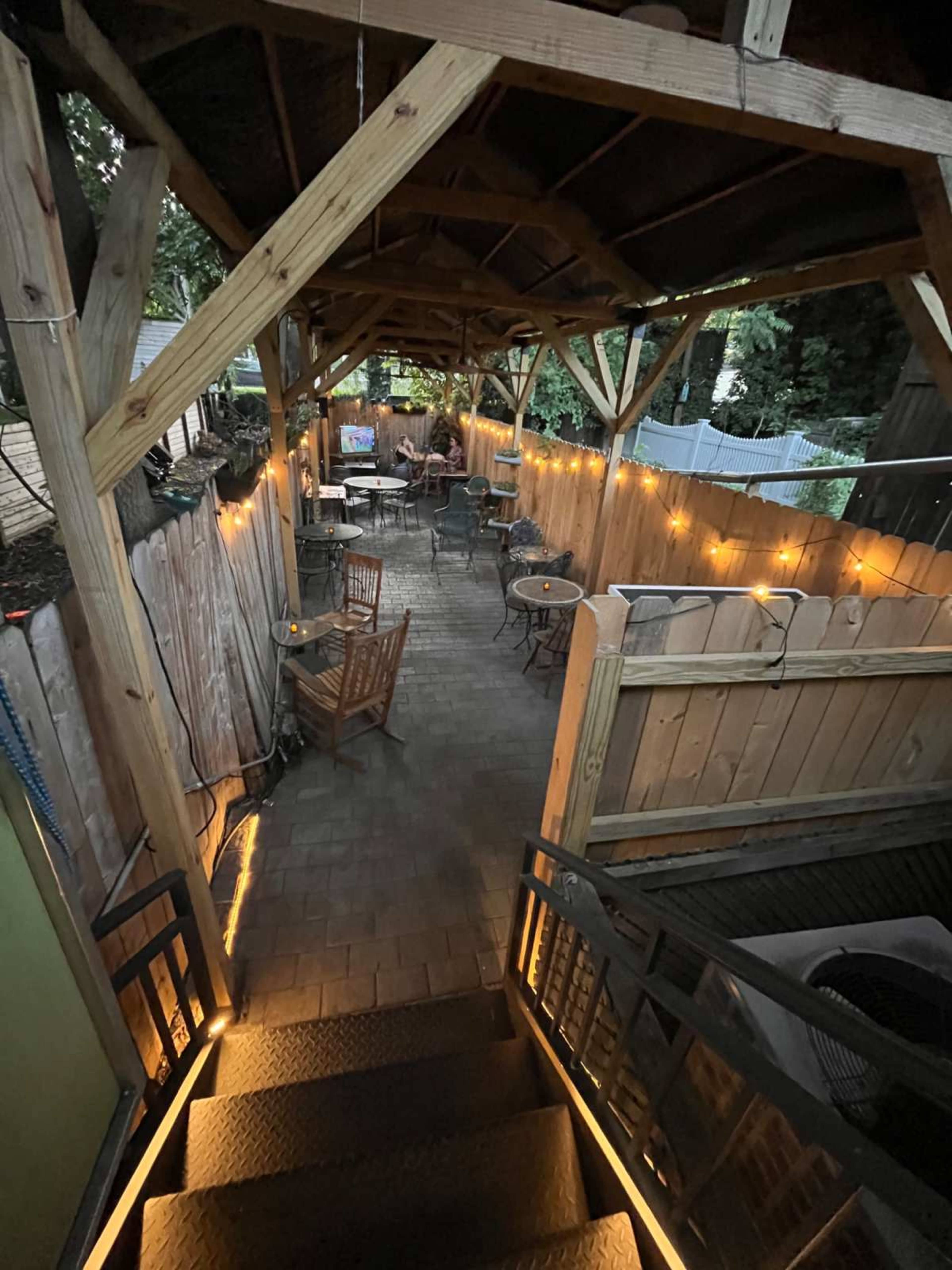 The image shows a patio area with wooden beams, stone flooring, and several tables and chairs illuminated by string lights along a wooden fence.