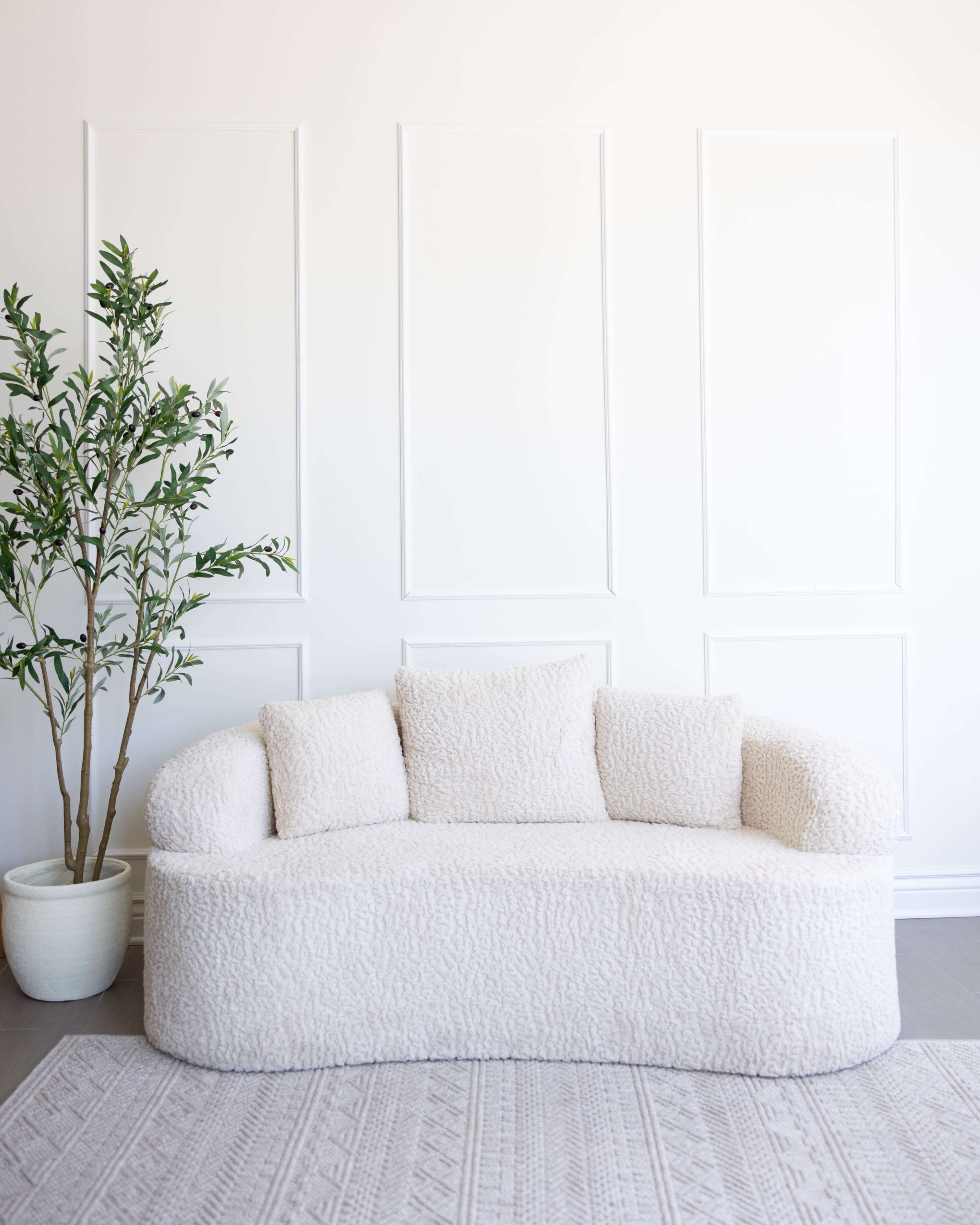 The image features a white, plush, curved sofa placed against a plain white wall, accompanied by a potted plant to the left.