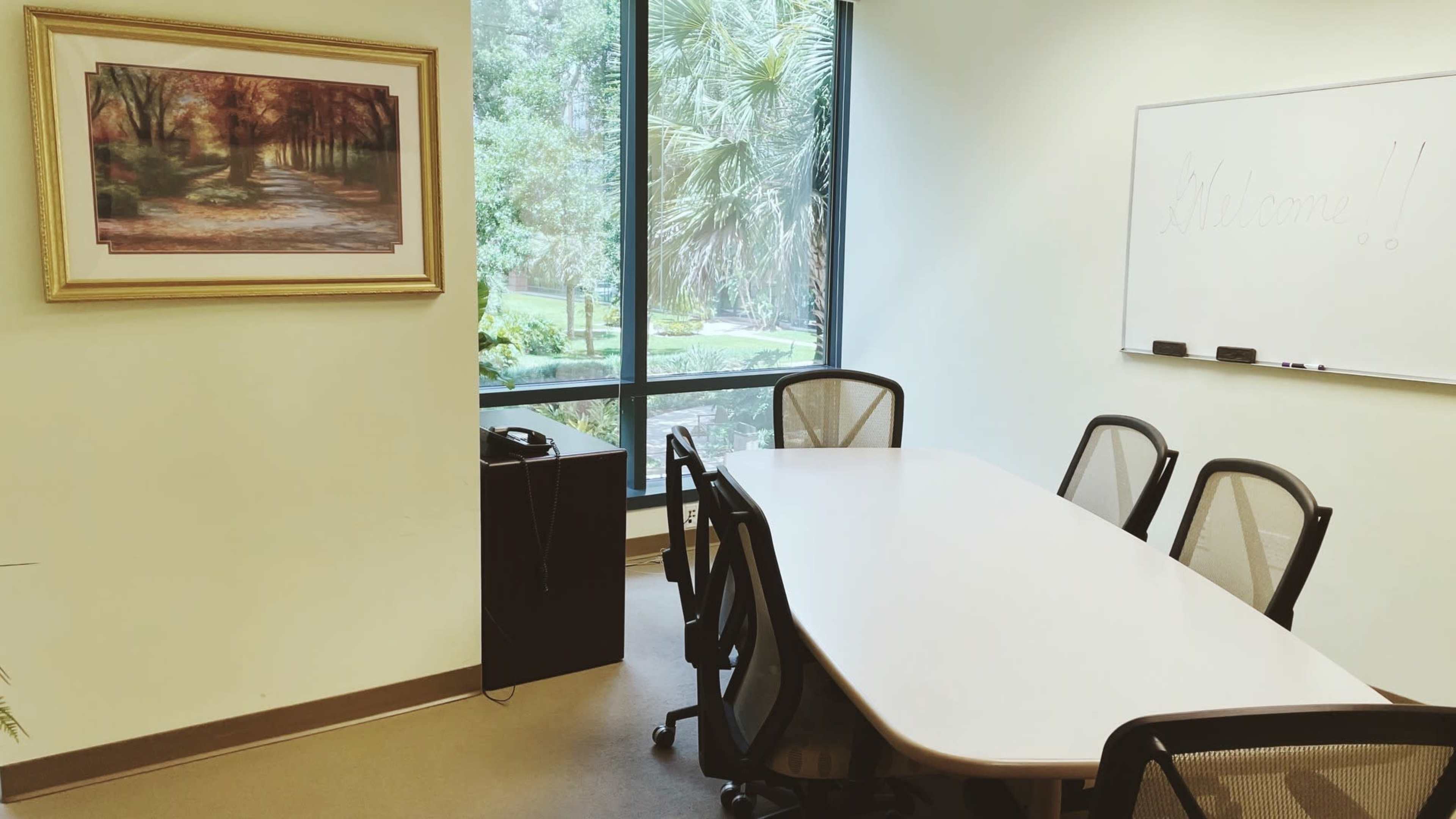 A conference room features a long table surrounded by chairs, a whiteboard, a framed landscape painting, and a window with a view of greenery.