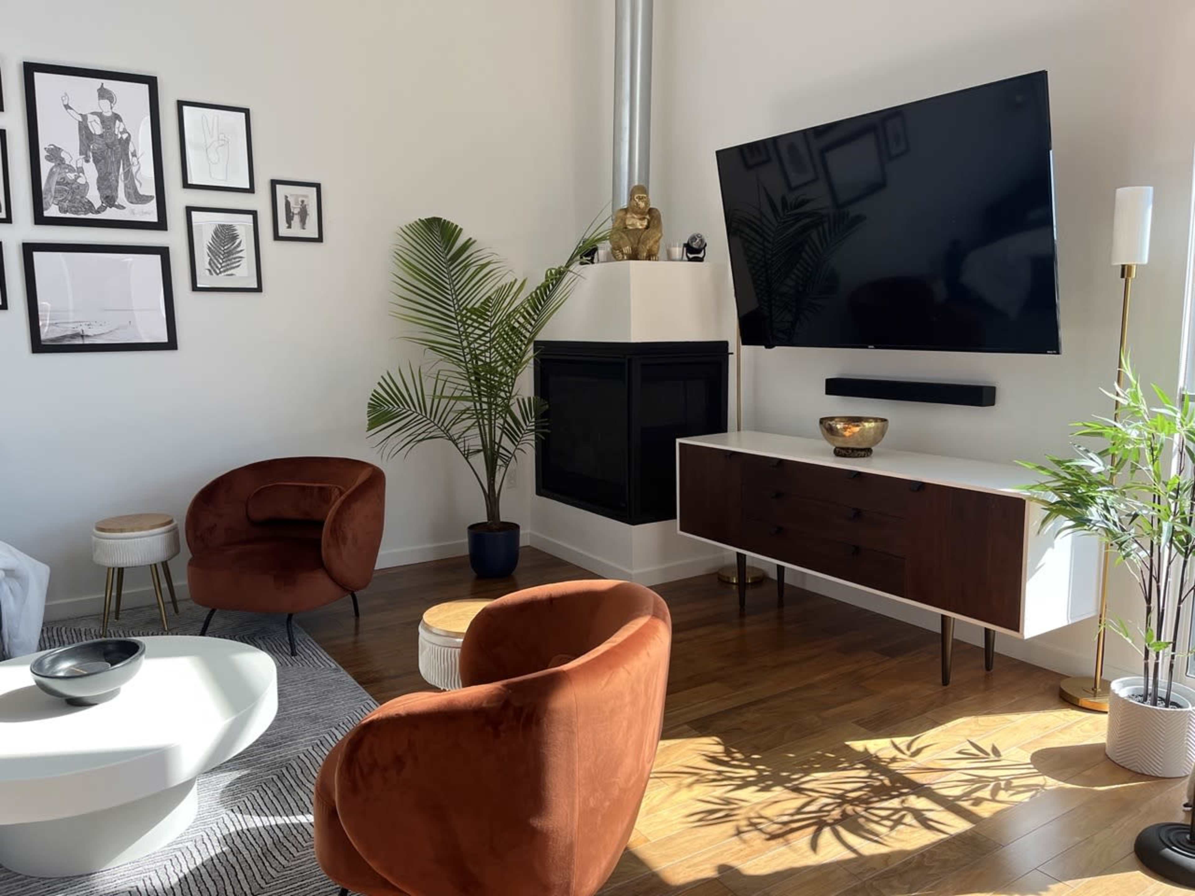 The room features a modern living area with two brown chairs, a white coffee table, a wall-mounted TV, and decorative plants.