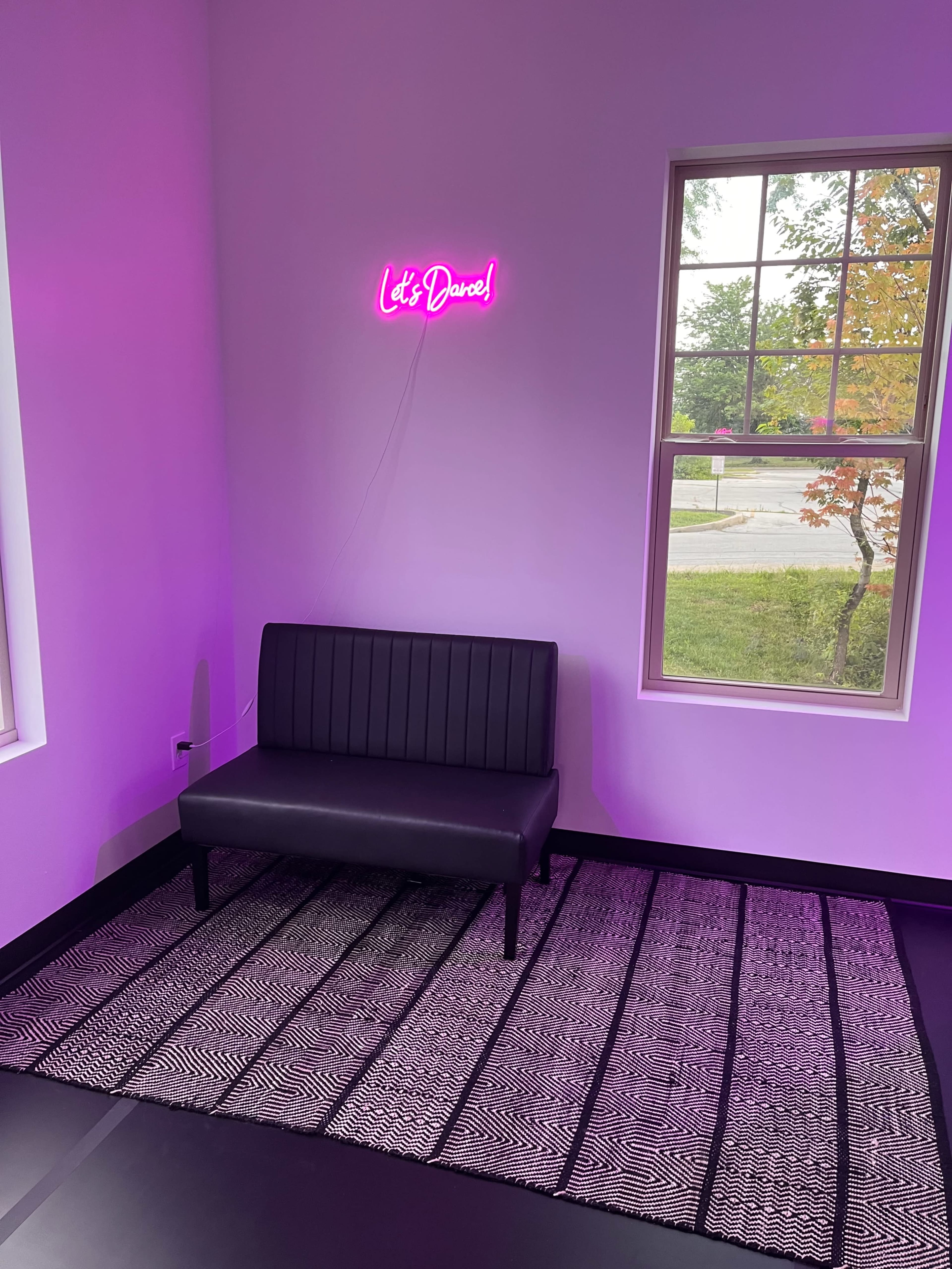A black bench sits against a pink-hued wall, illuminated by a neon sign that says "Let's Dance," while a patterned rug lies on the floor beneath it.