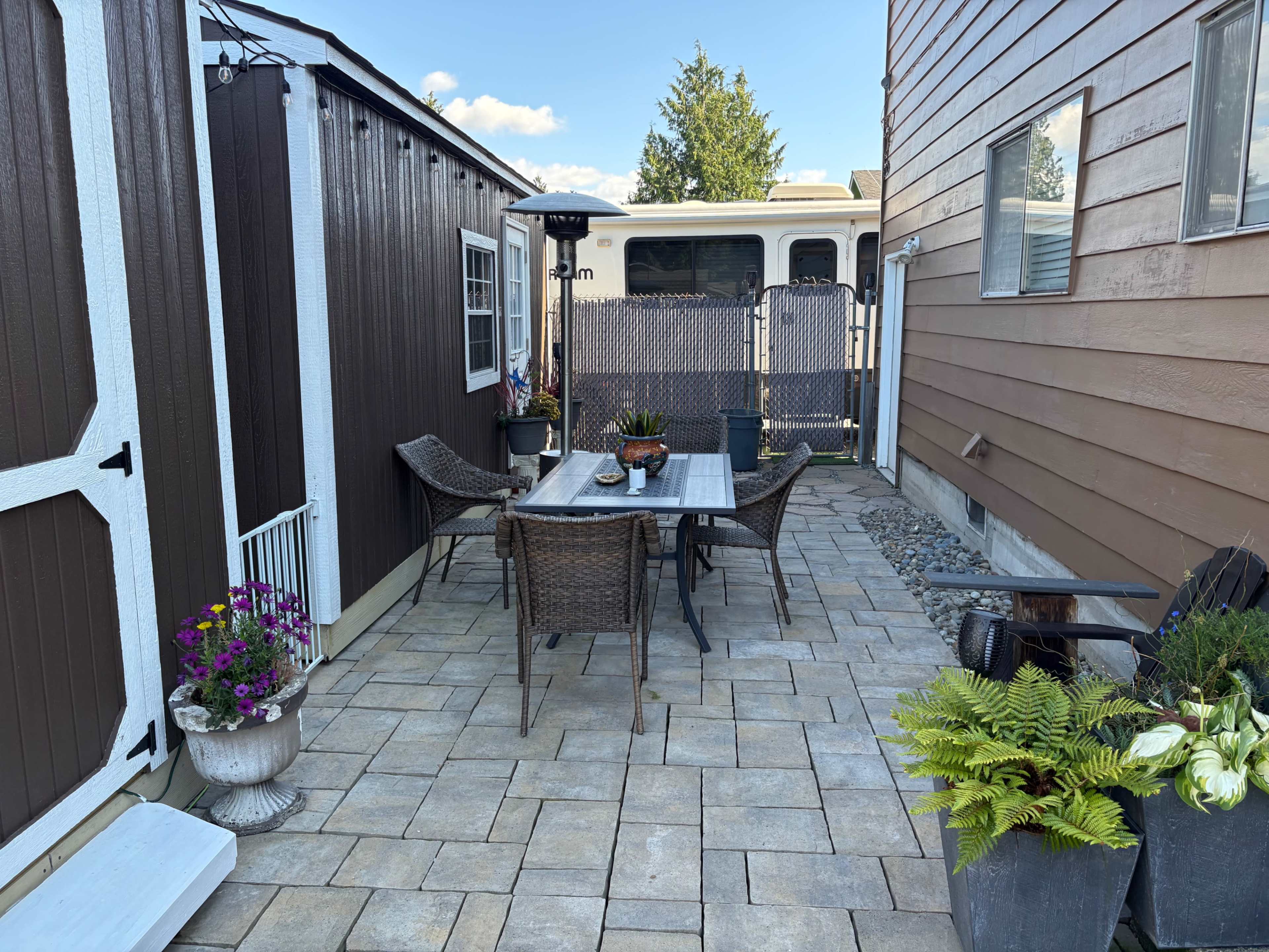 The image shows a backyard patio area with a table and four chairs, surrounded by decorative plants and a storage shed, with a trailer partially visible in the background.