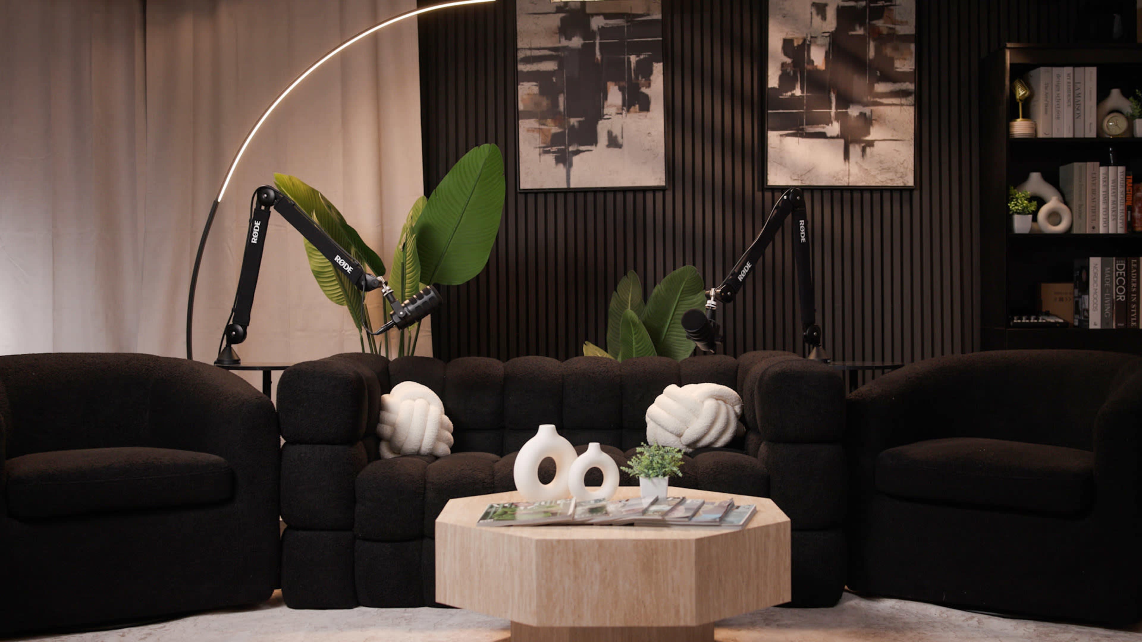 The scene features a modern living room with a black upholstered sofa, a round wooden coffee table adorned with decorative items, and large green plants alongside abstract wall art.