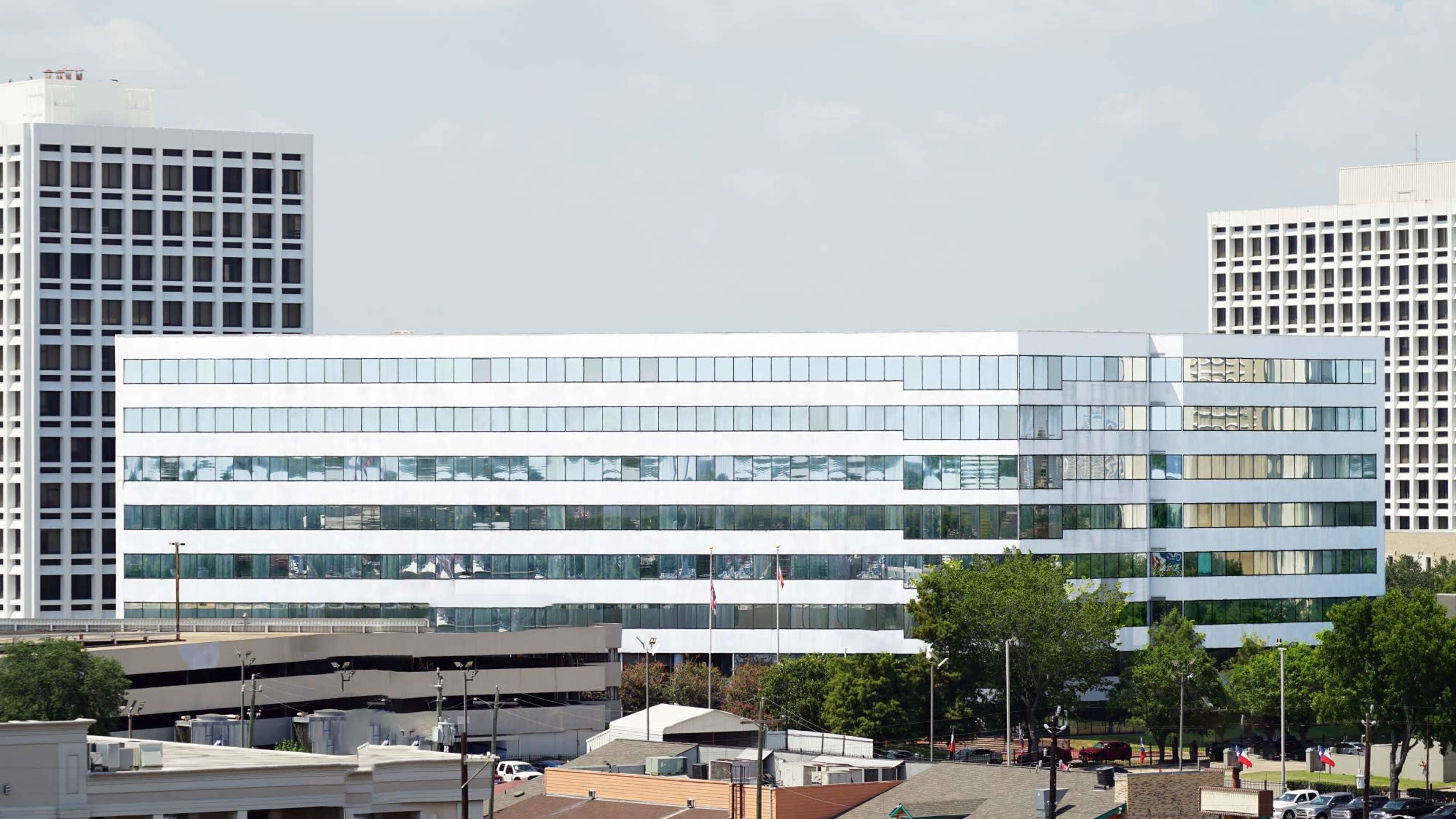A modern glass-covered office building flanked by two taller structures, with a parking area and trees in the foreground.