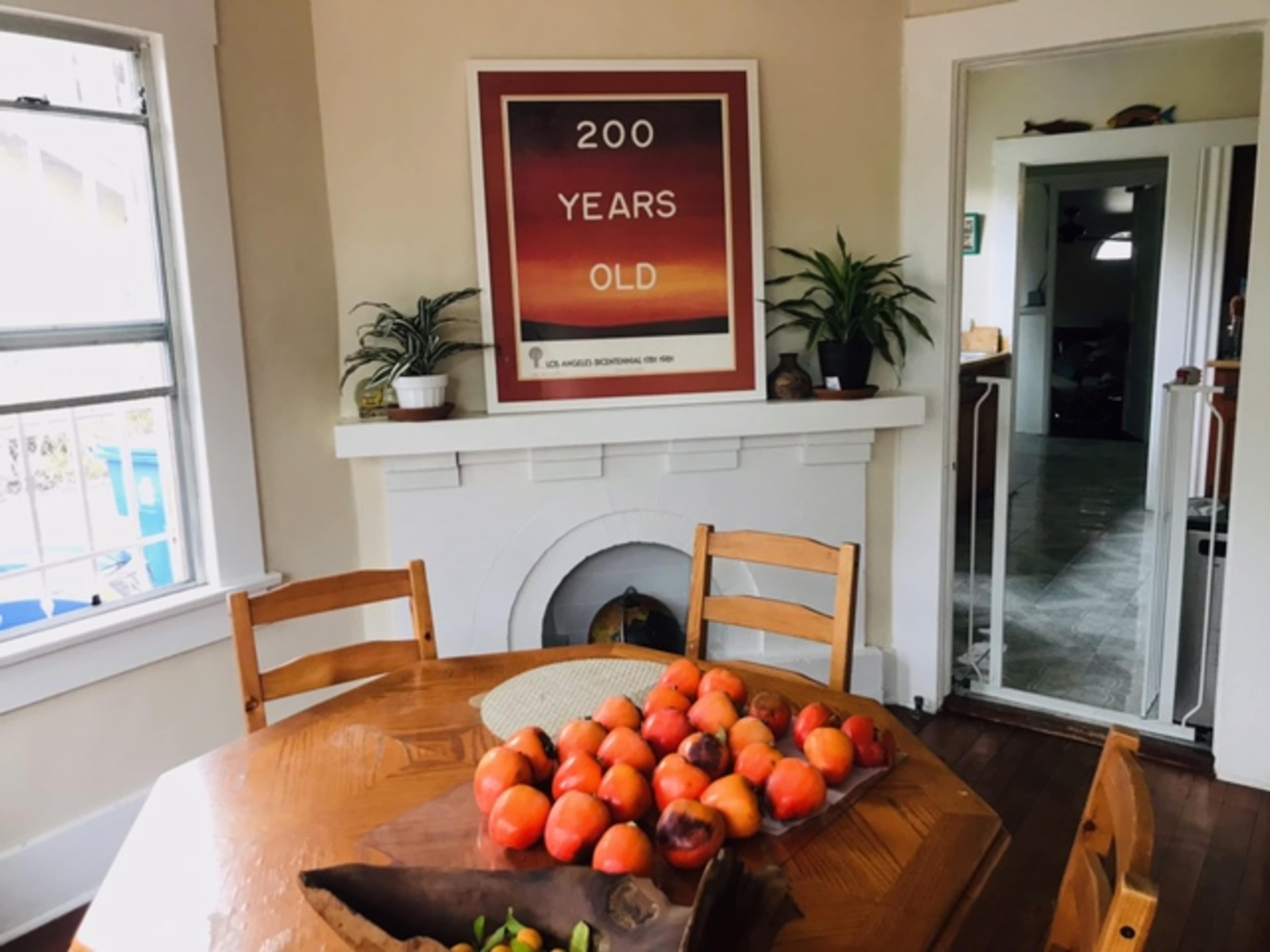 A wooden dining table with a bowl of persimmons is set in a room featuring a poster that reads "200 YEARS OLD" on the wall.