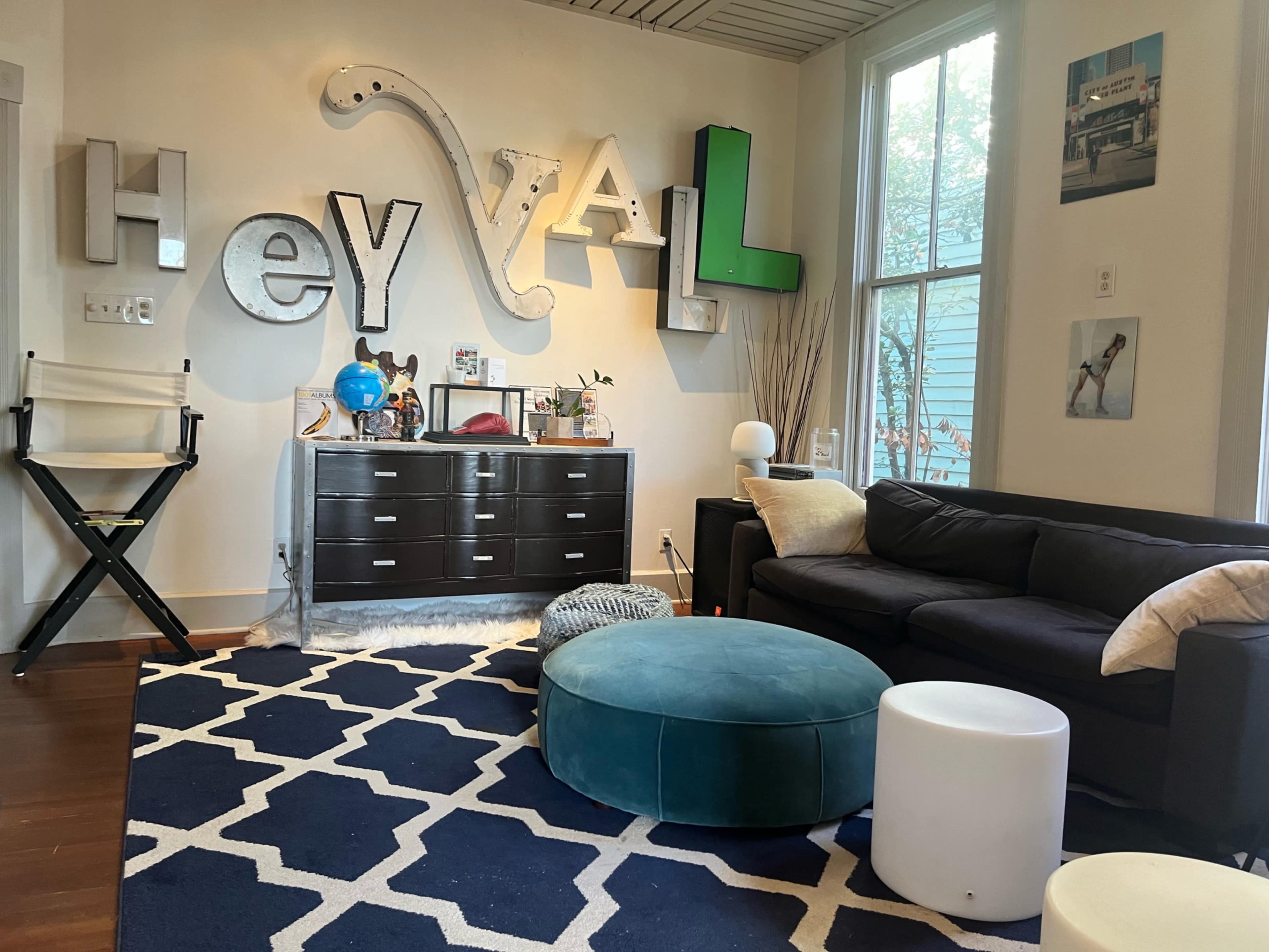 The scene features a living room with a dark sofa, a blue pouf, a white side table, and large decorative letters spelling "Hey Y'all" on the wall.