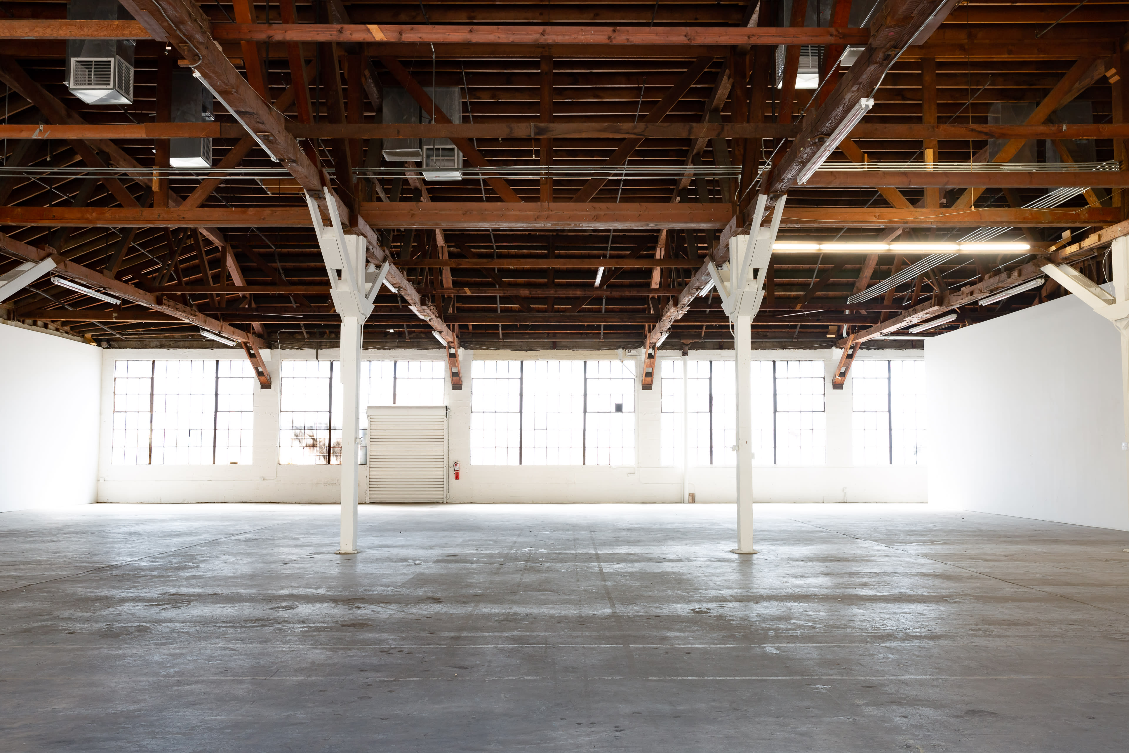 The image shows a spacious, empty industrial room with wooden beams and large windows along one wall.