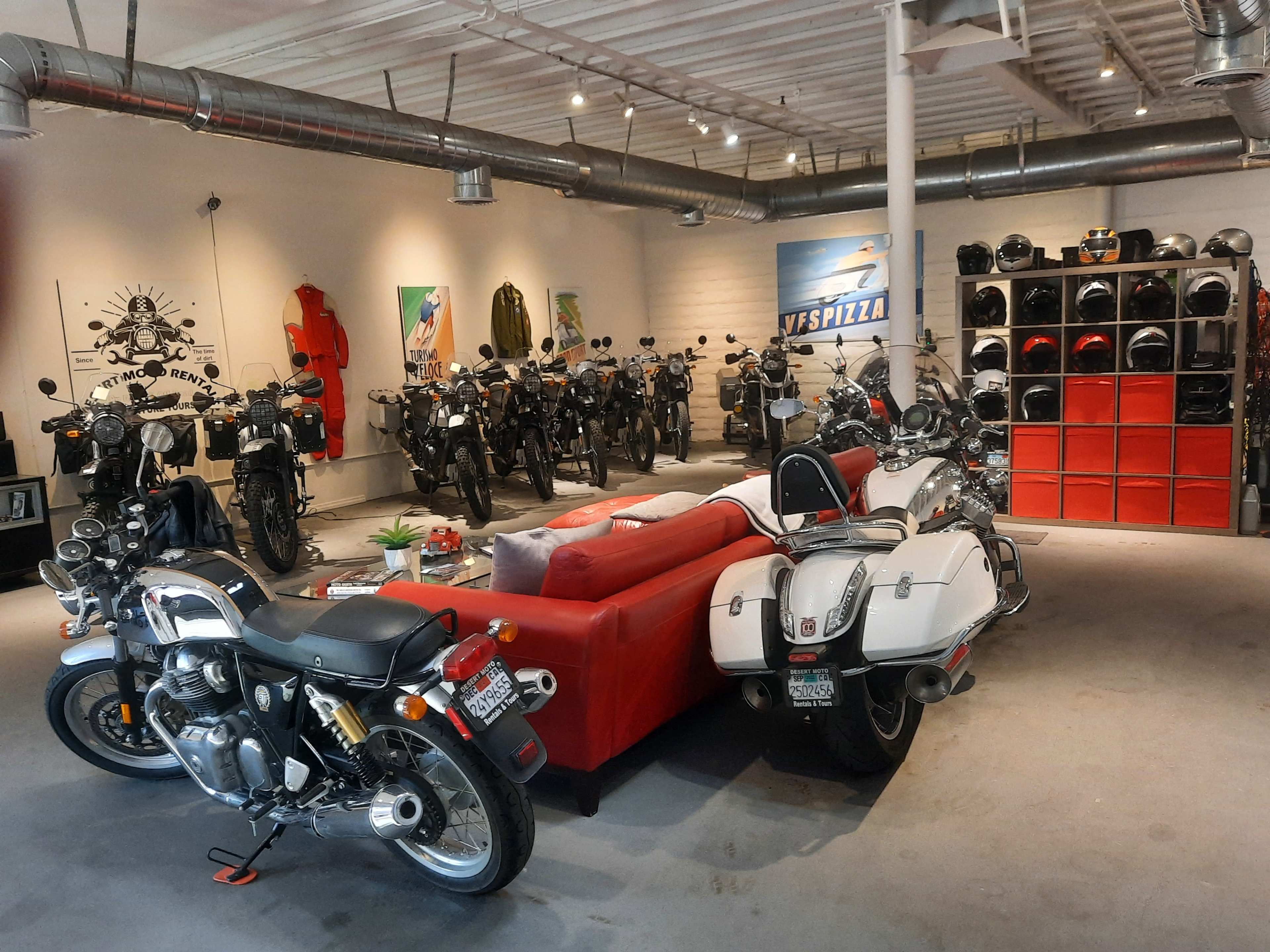 A spacious motorcycle showroom featuring several bikes arranged along the walls, with a red sofa in the foreground and a collection of helmets displayed on shelves.