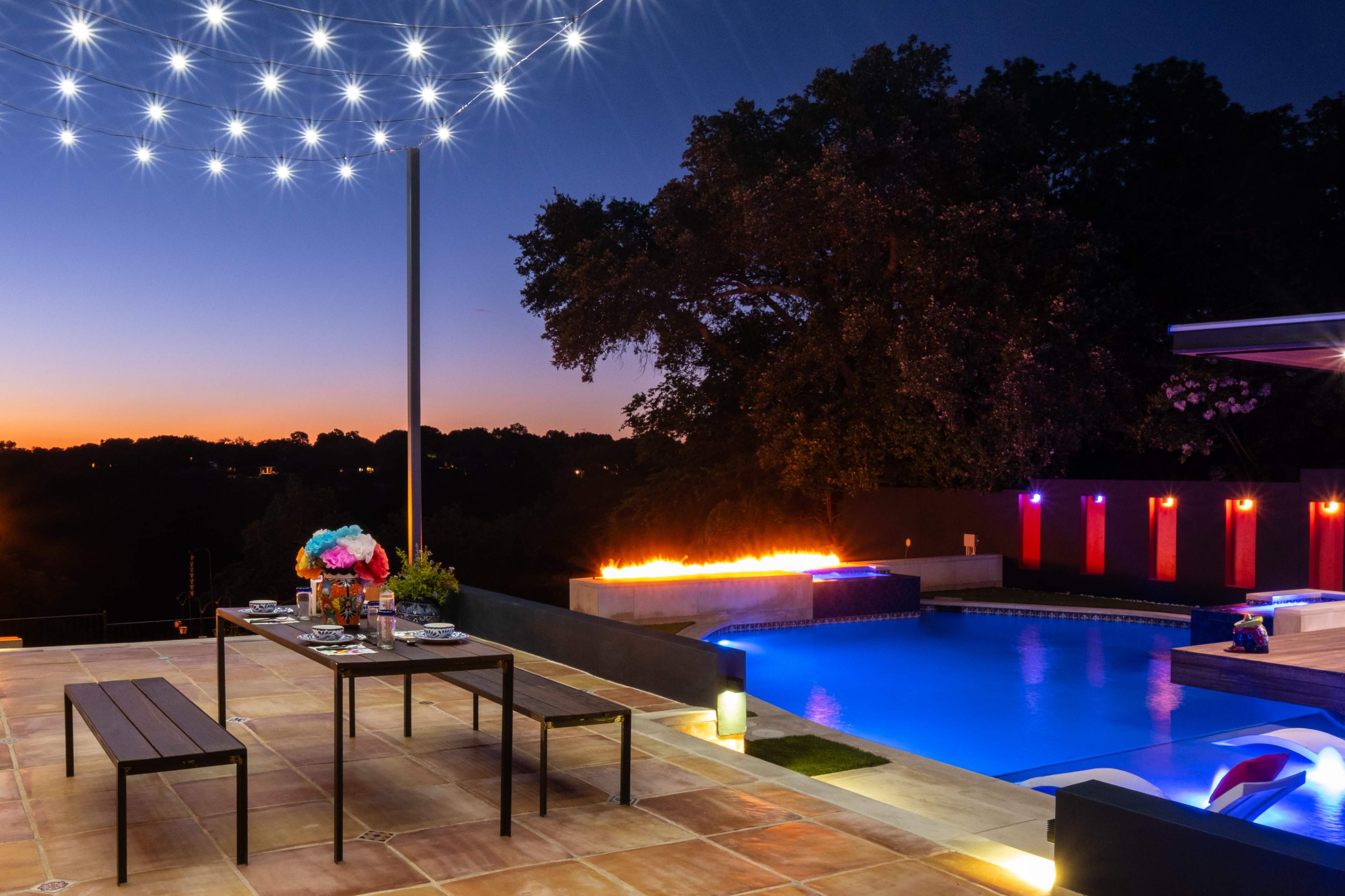 A patio area features a dining table set near a brightly lit swimming pool with a fire feature, surrounded by trees at dusk.