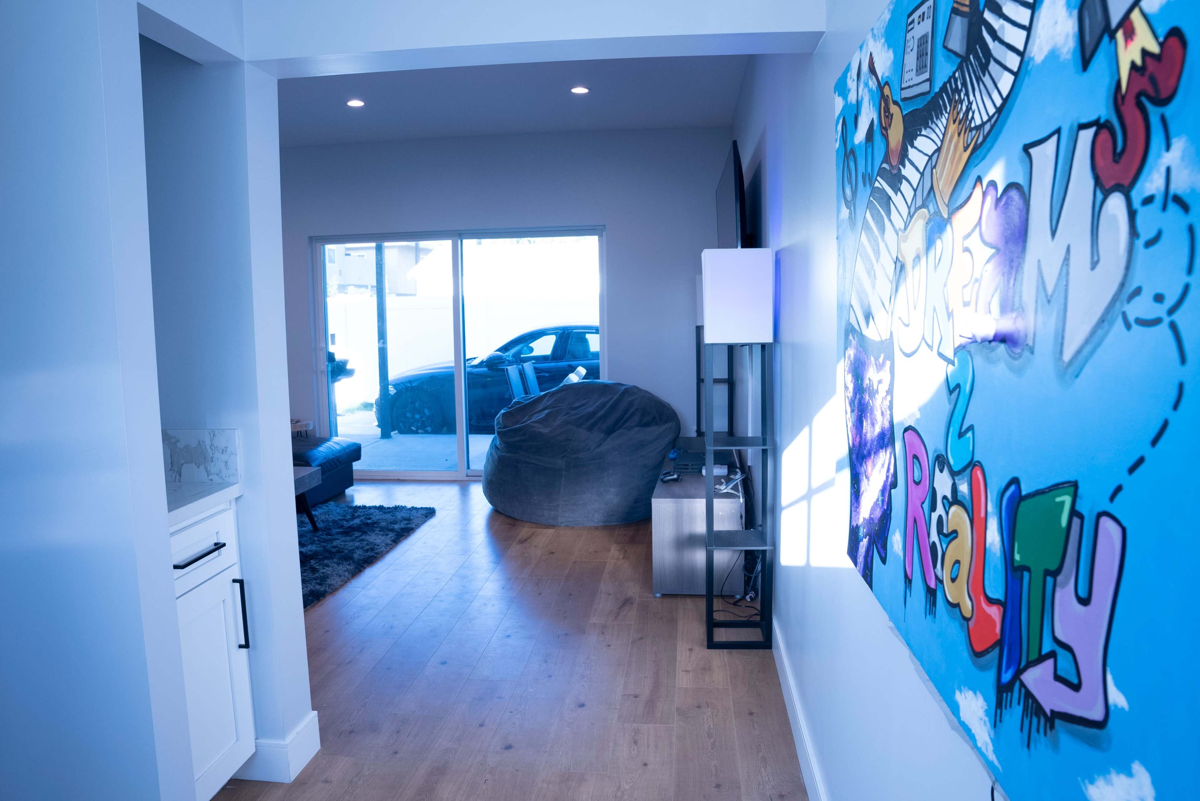 A brightly lit room features a colorful mural on one wall, with a plush bean bag in the corner and a view of a car parked outside through sliding glass doors.