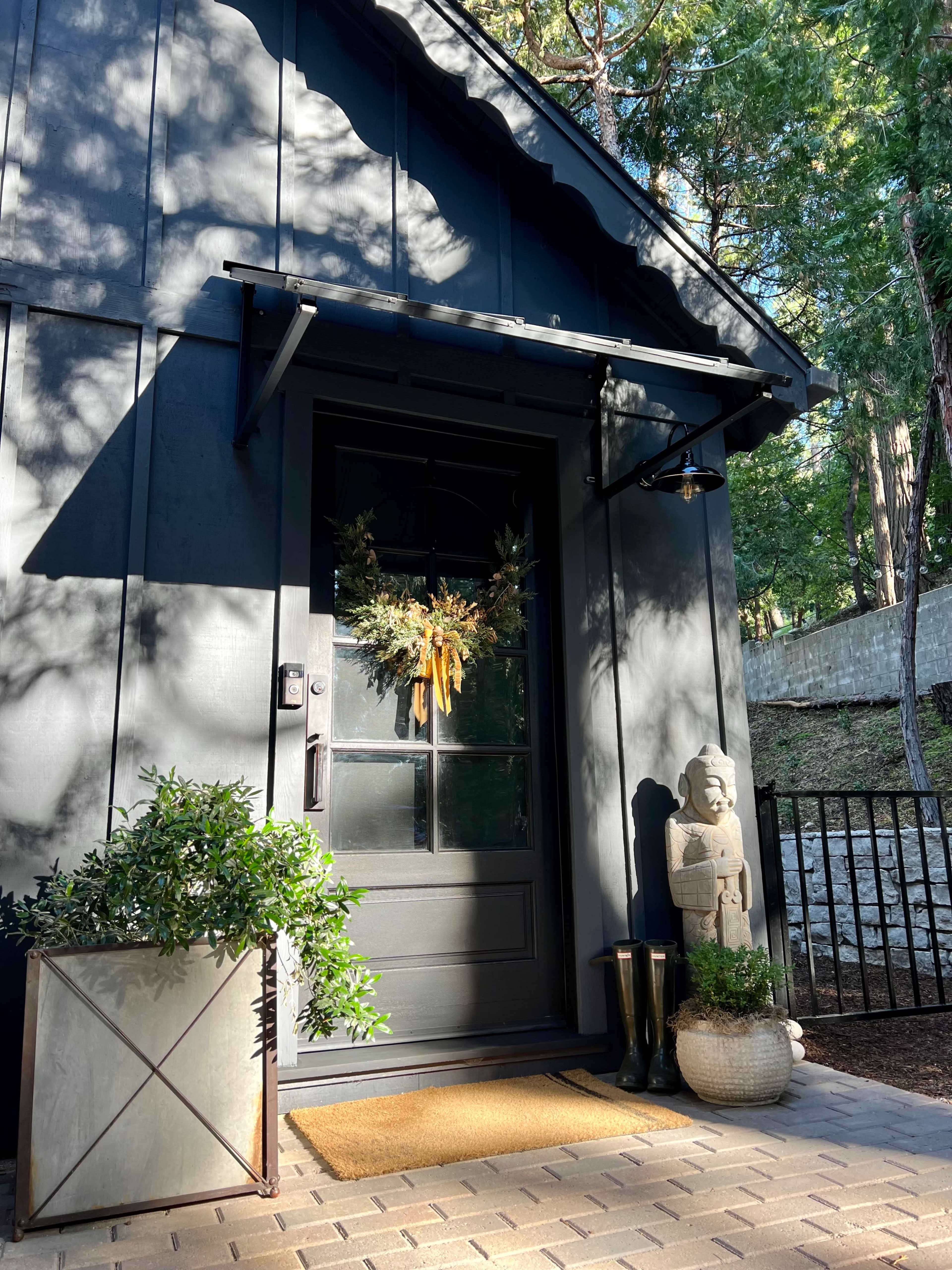 A dark exterior door adorned with a seasonal wreath, flanked by potted plants and decorative items, set against a backdrop of greenery.