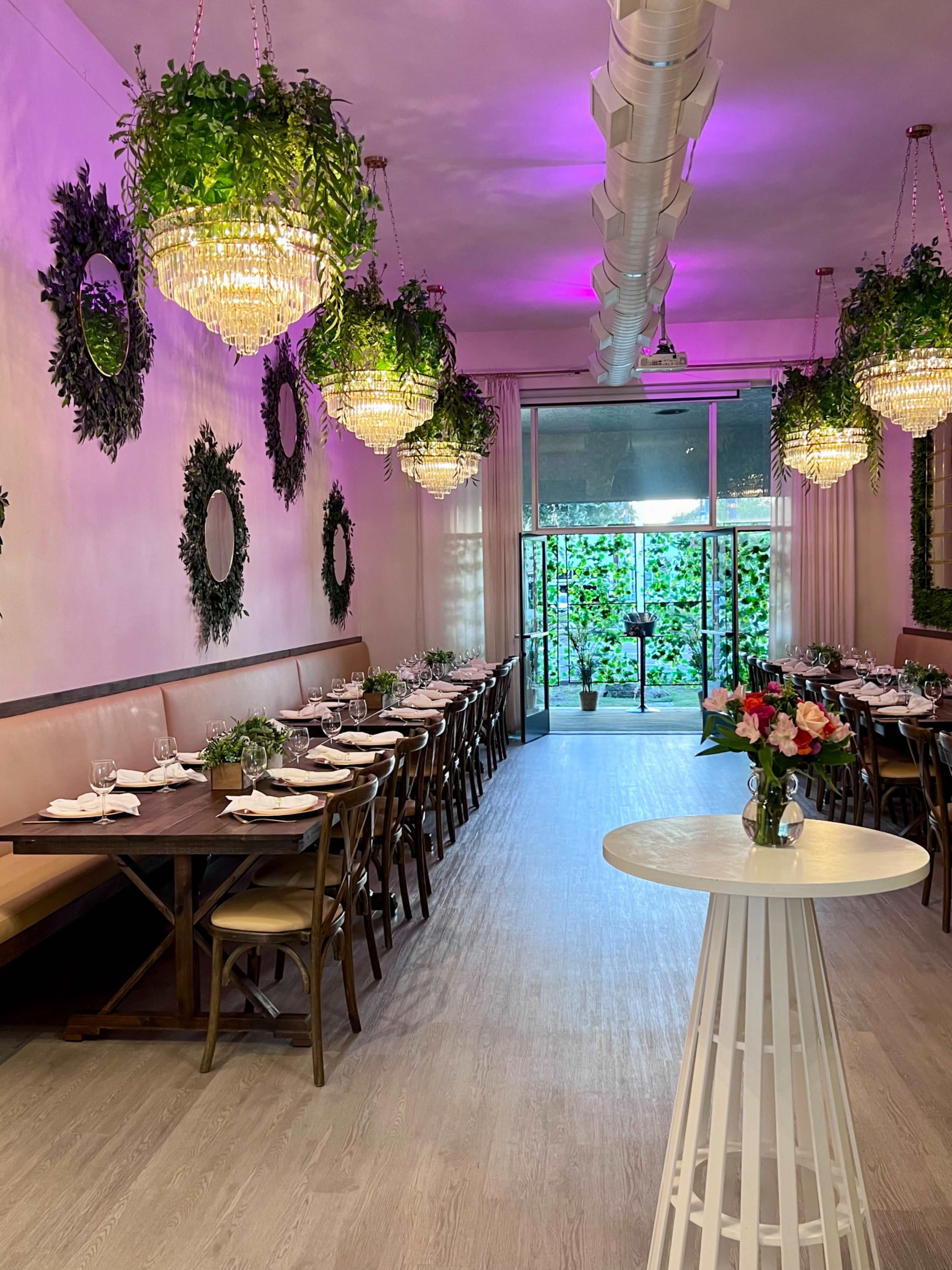 The image shows a modern restaurant interior with wooden tables set for dining, adorned with potted plants and chandeliers, and a glass door leading to an outdoor garden.