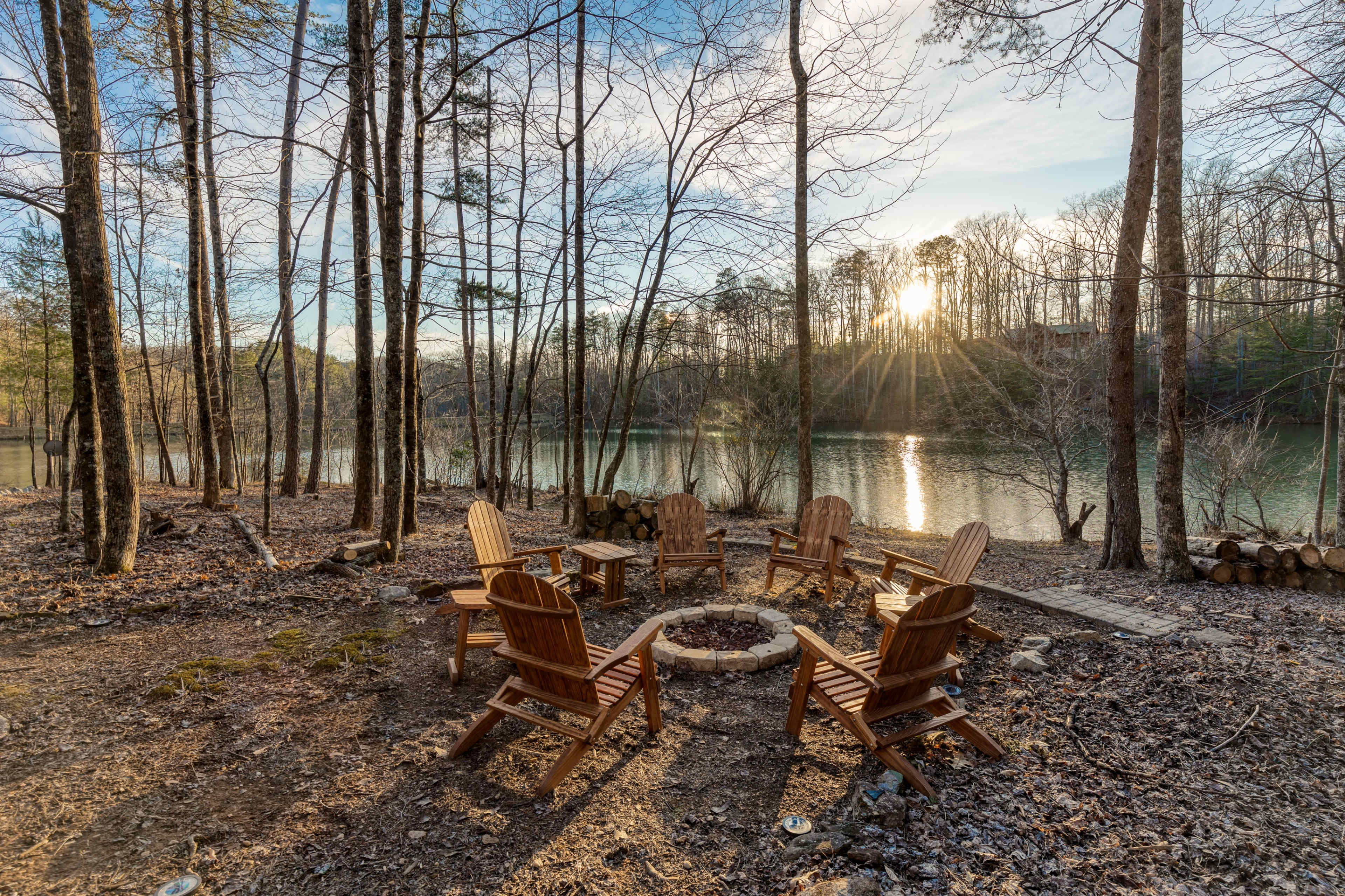 A circle of wooden chairs surrounds a fire pit by a lake, framed by trees reflecting the setting sun.