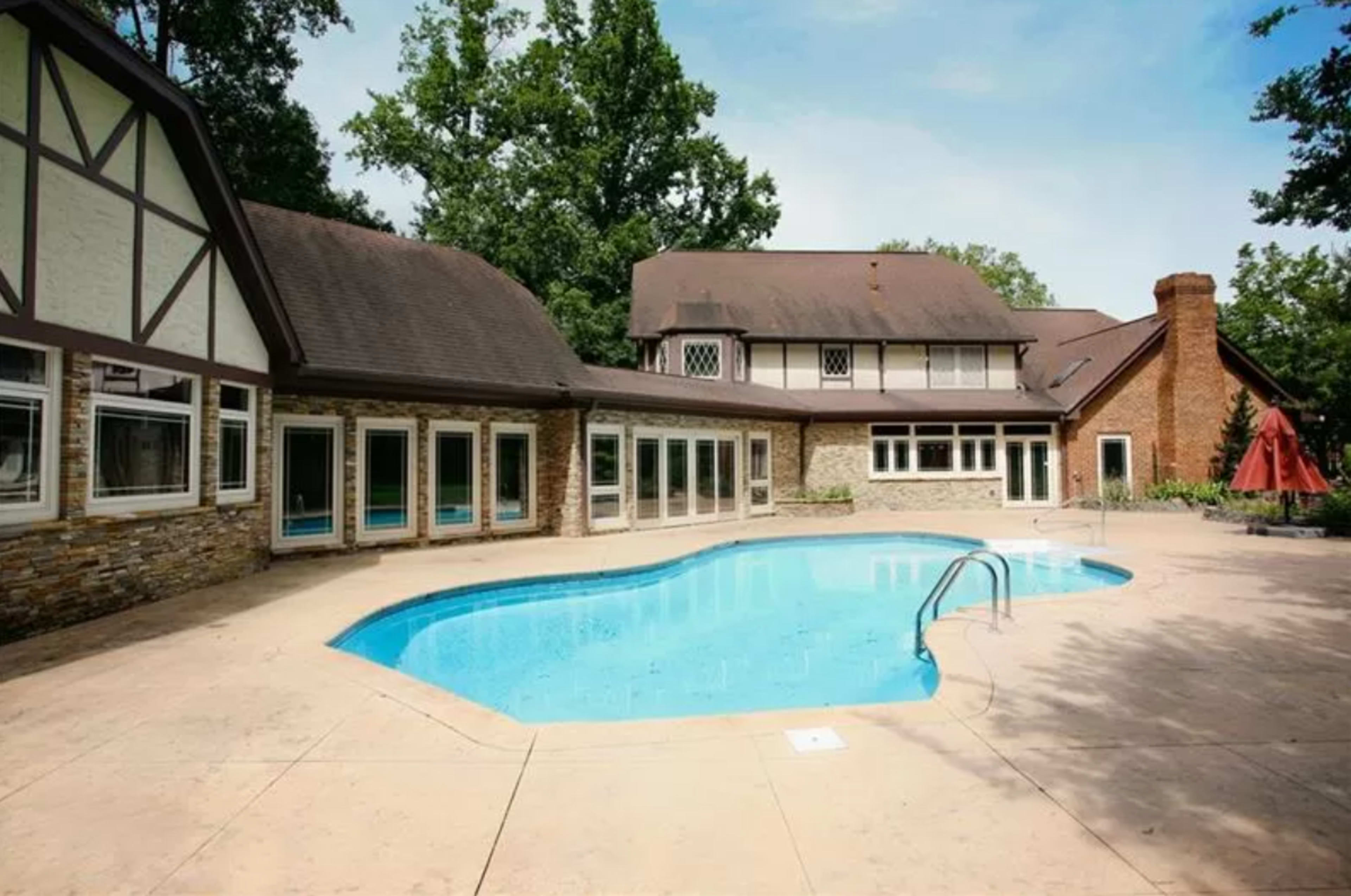 A large, rectangular swimming pool is surrounded by a stone and tile patio, with a two-story house featuring a mix of brick and timber architecture in the background.