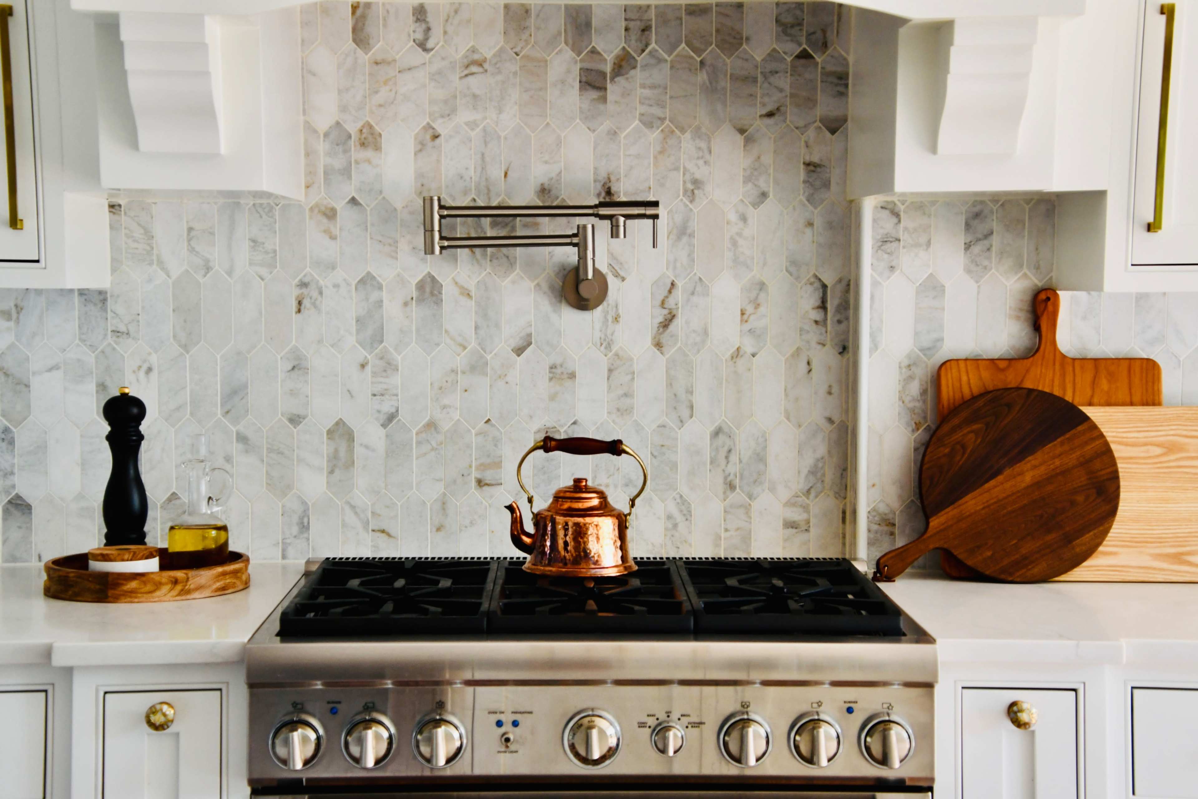 A copper kettle sits on a stainless steel stove against a backdrop of hexagonal marble tiles, with wooden cutting boards and a pepper mill nearby.