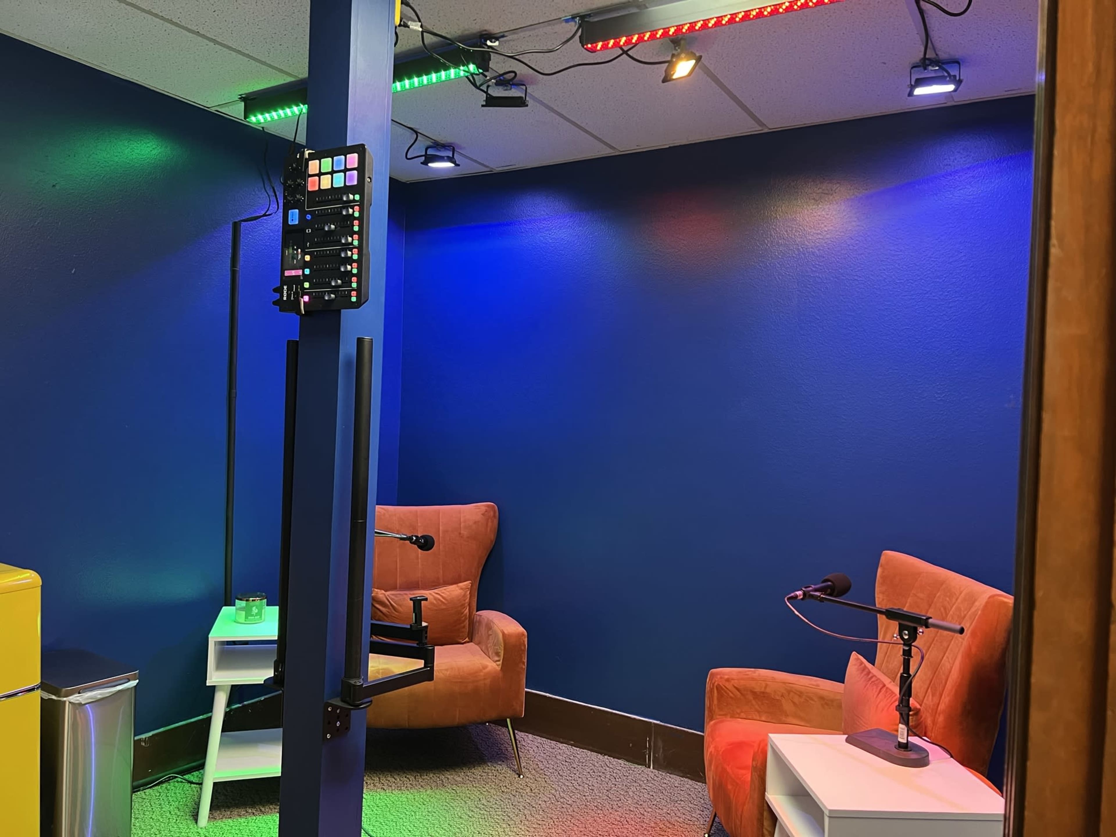 The image shows a small room with two orange chairs, a white side table, a microphone on a stand, and colored lights illuminating the blue walls.