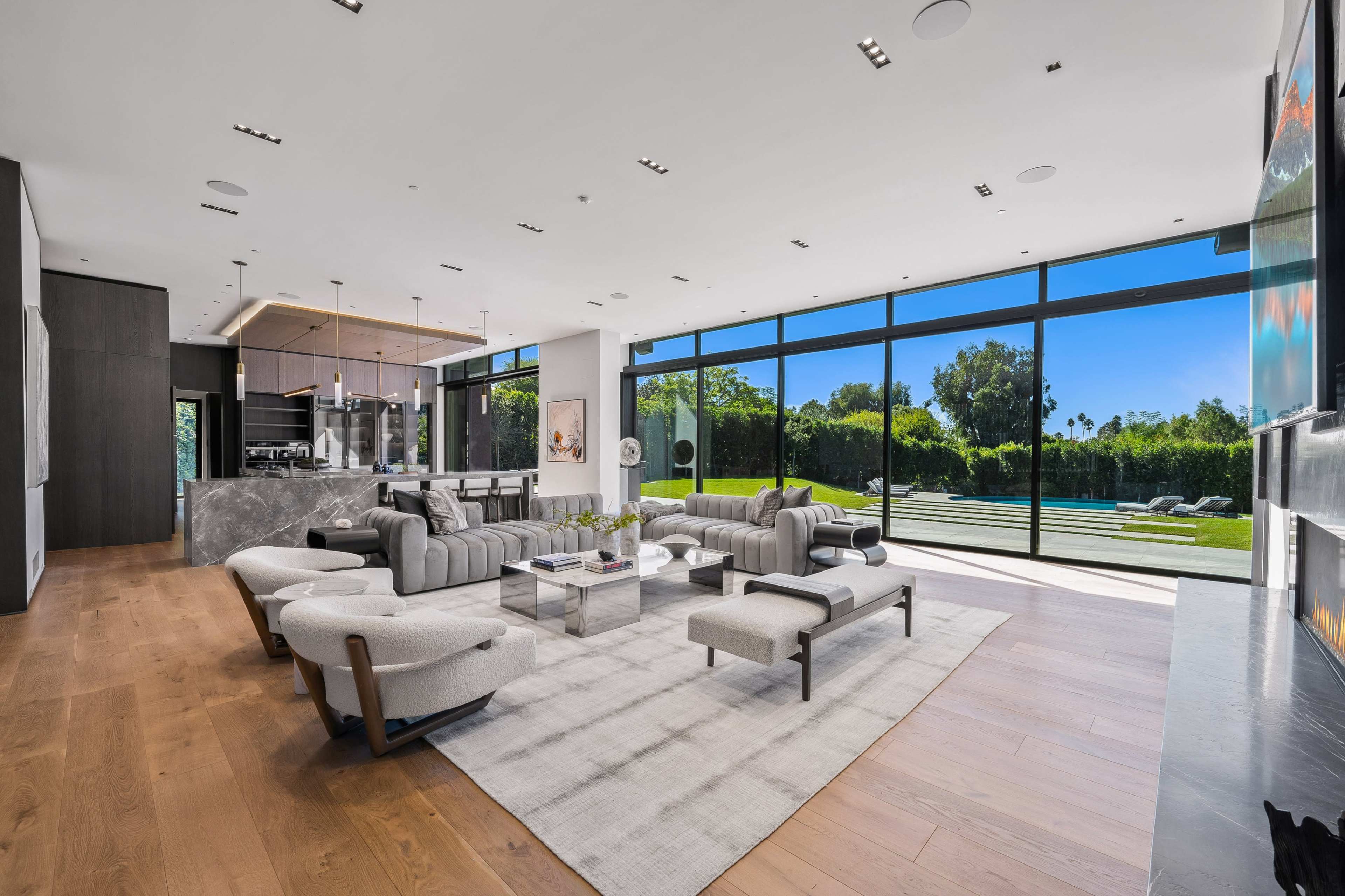 A modern living room features a spacious layout with large windows overlooking a garden, sleek furniture, and wooden flooring.
