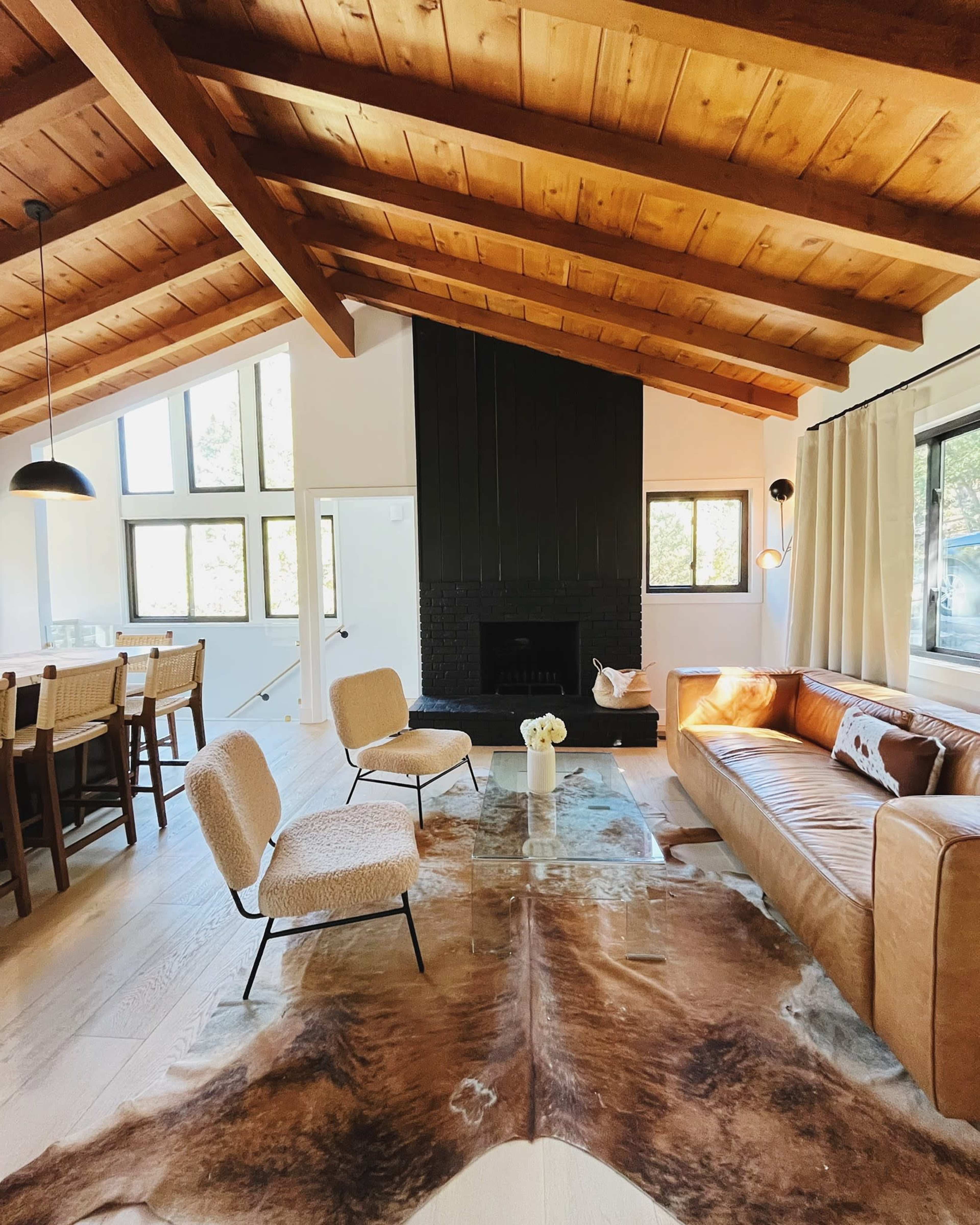 The living space features a brown leather sofa, two upholstered chairs, a rustic cowhide rug, and a fireplace, with large windows allowing natural light to fill the room.