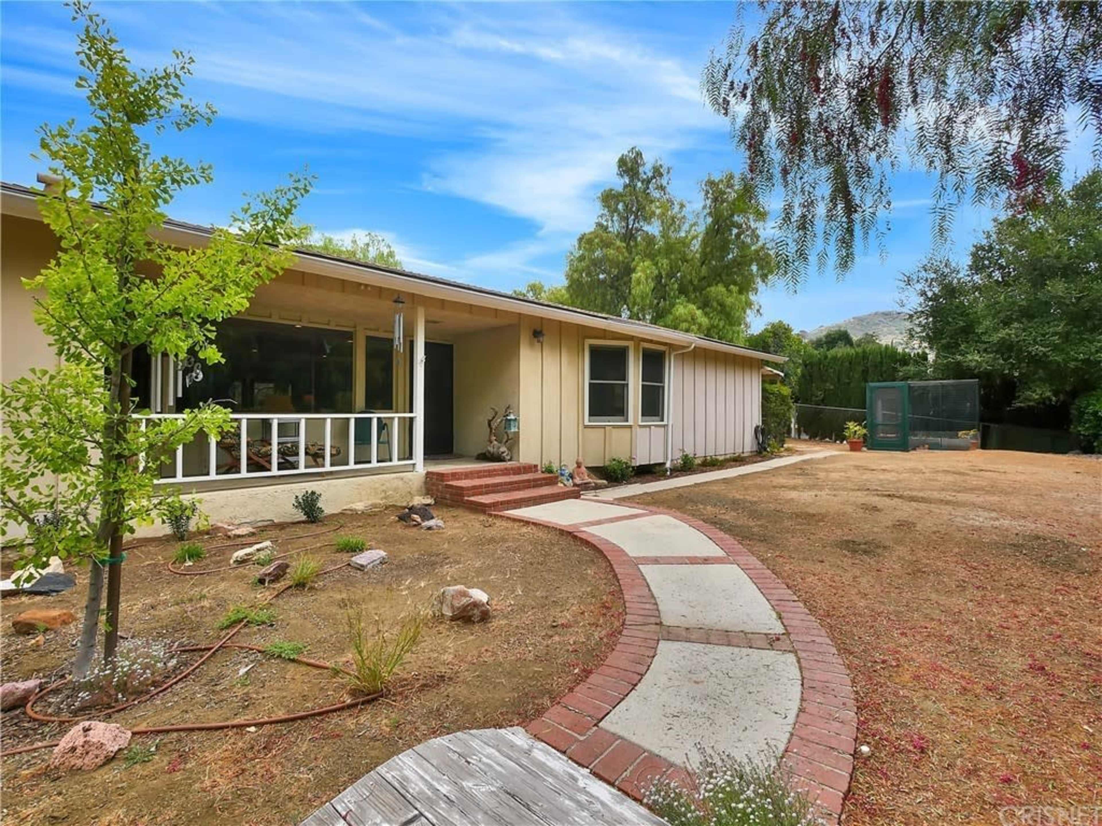A single-story house with a brick walkway leads to a front porch set amid a yard with sparse vegetation and a fenced area in the background.