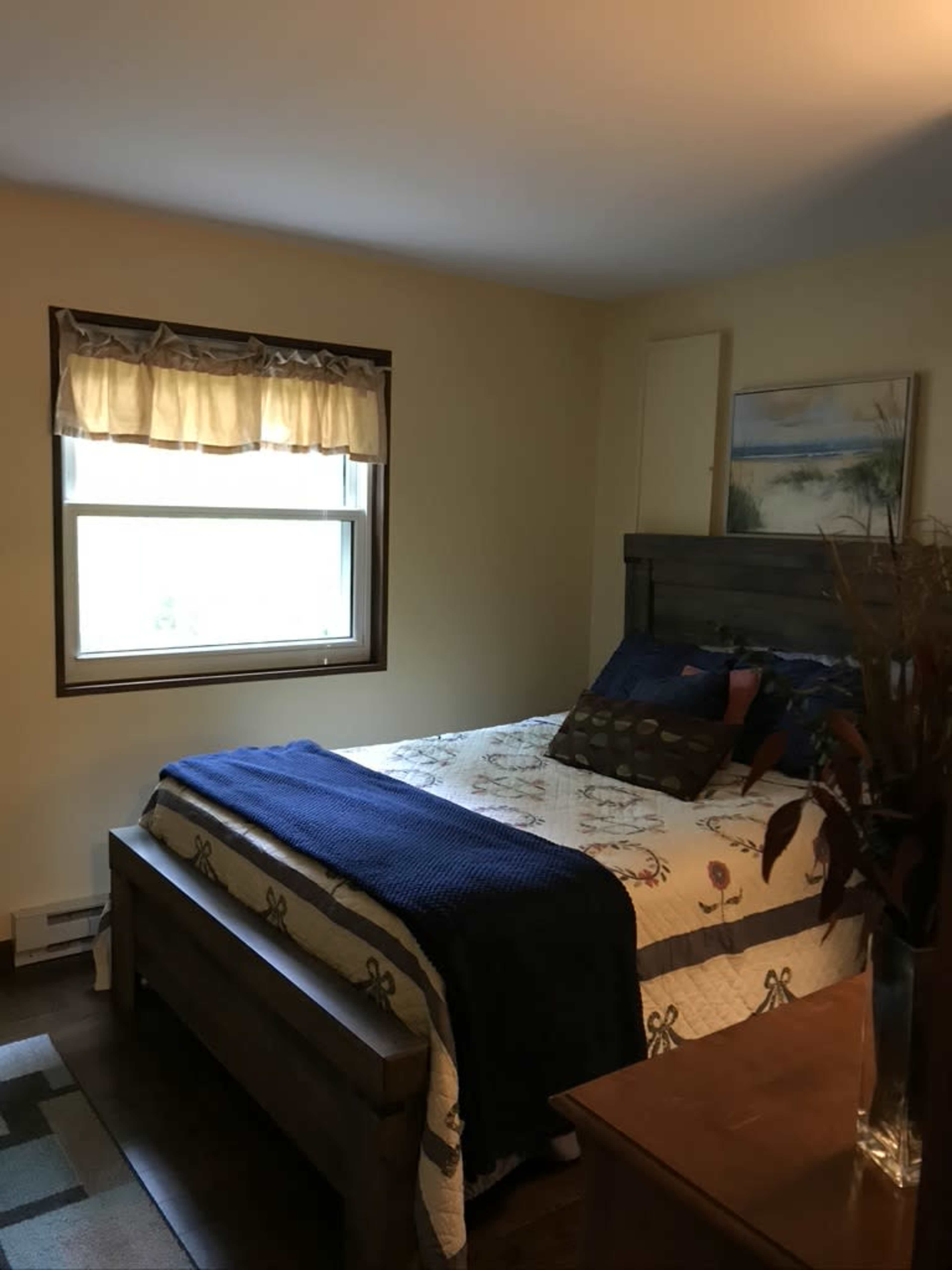 A neatly arranged bedroom features a bed with a quilt, a window with curtains, and a painting on the wall.