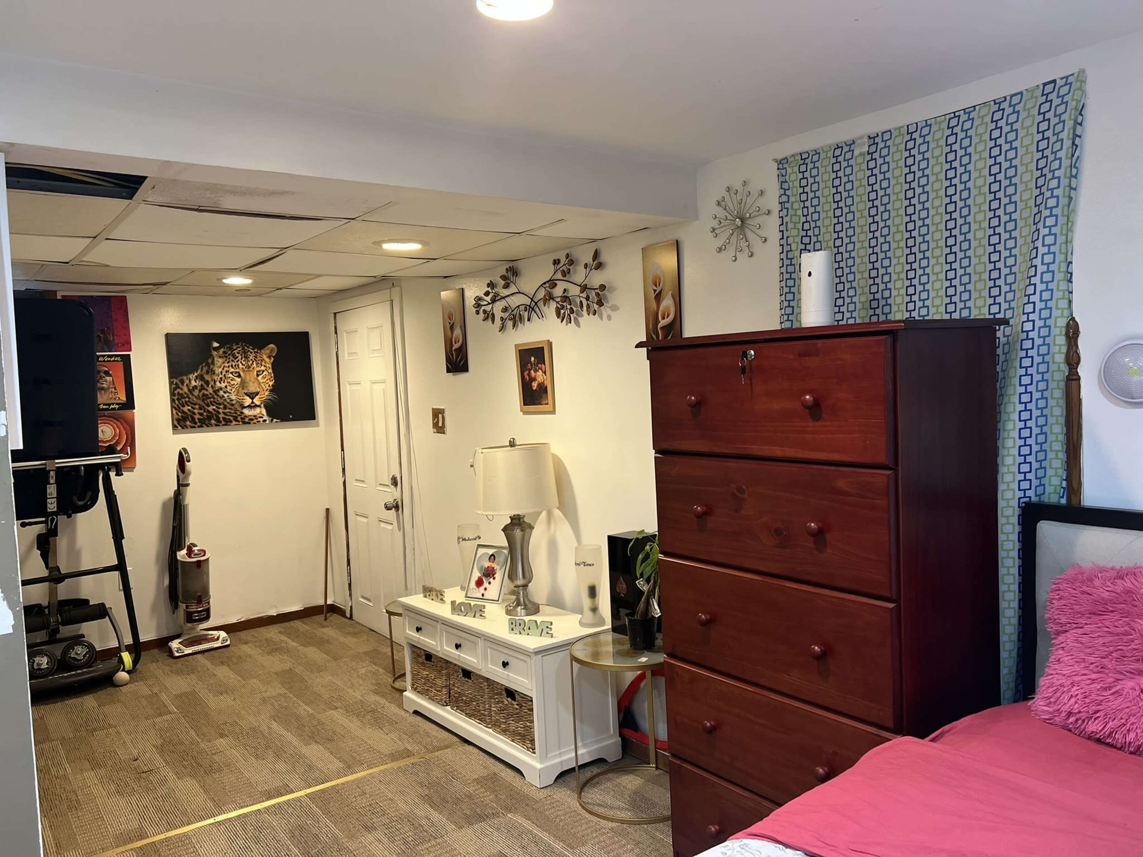 A bedroom featuring a wooden dresser, a nightstand with a lamp, and wall art, with a vacuum cleaner in the corner and a bed against one wall.