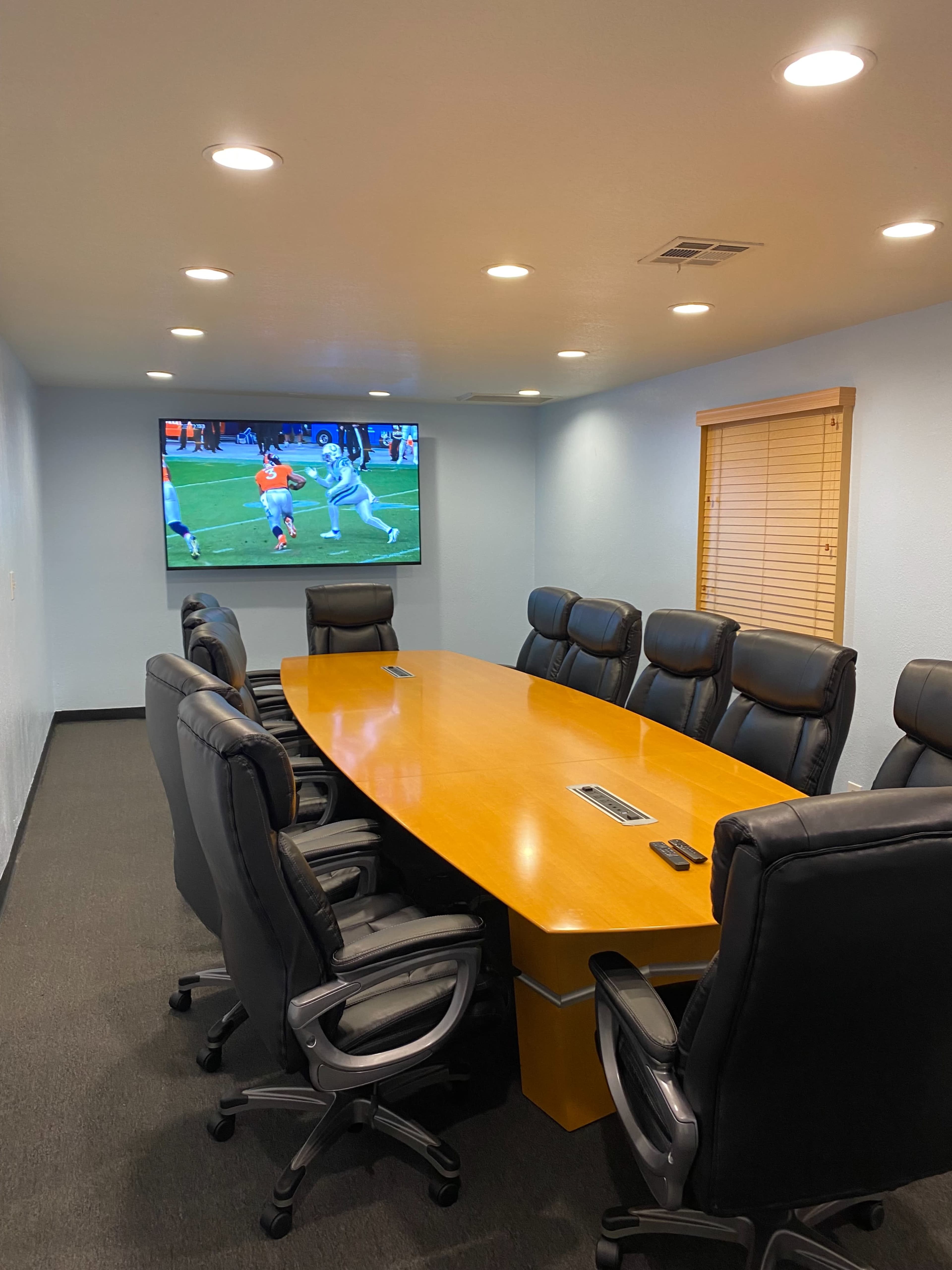 A conference room features a long wooden table surrounded by black leather chairs, with a television displaying a sports event on the wall.