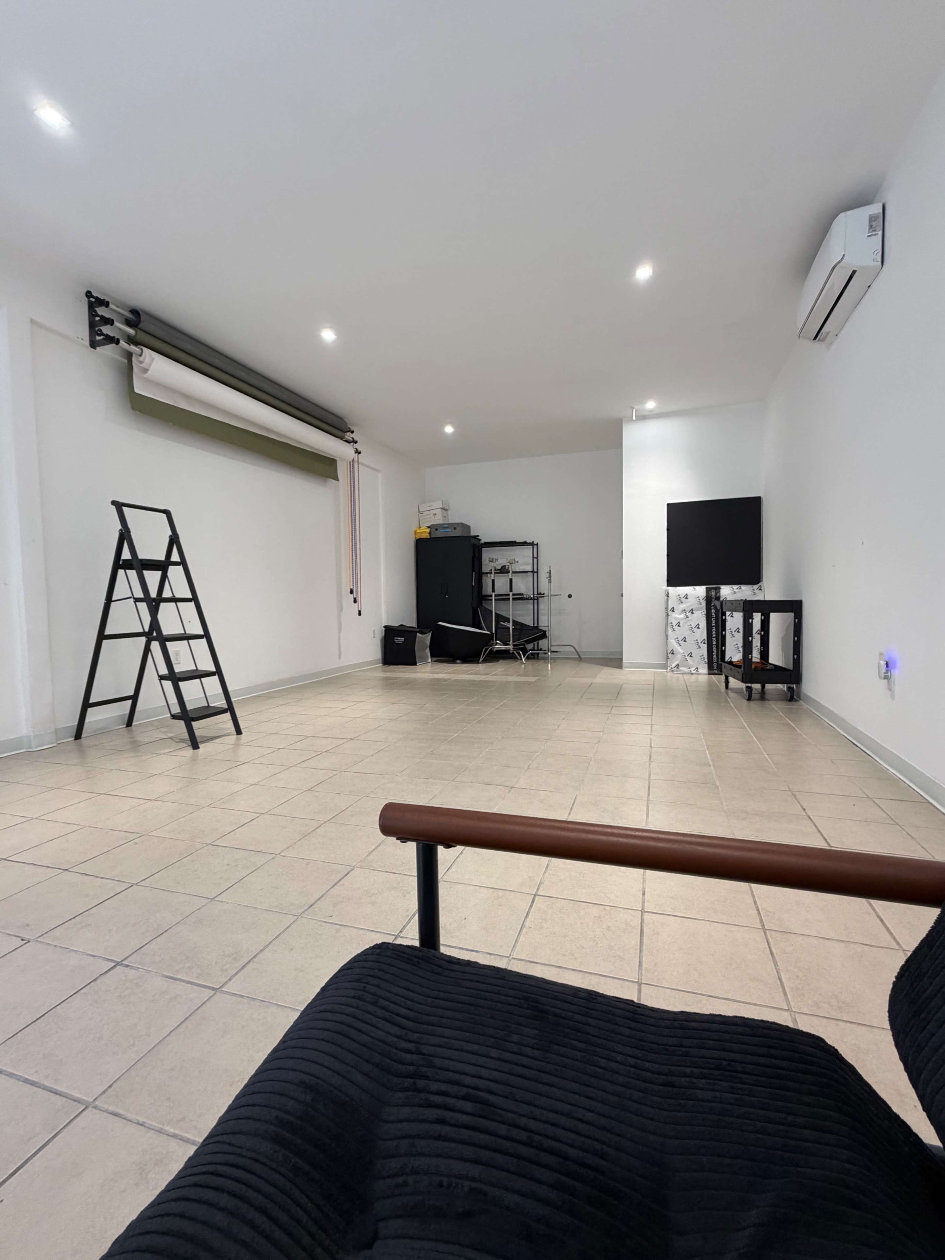 The image shows a spacious room with tiled floors, a black chair in the foreground, a ladder, and a television on a stand against the wall.
