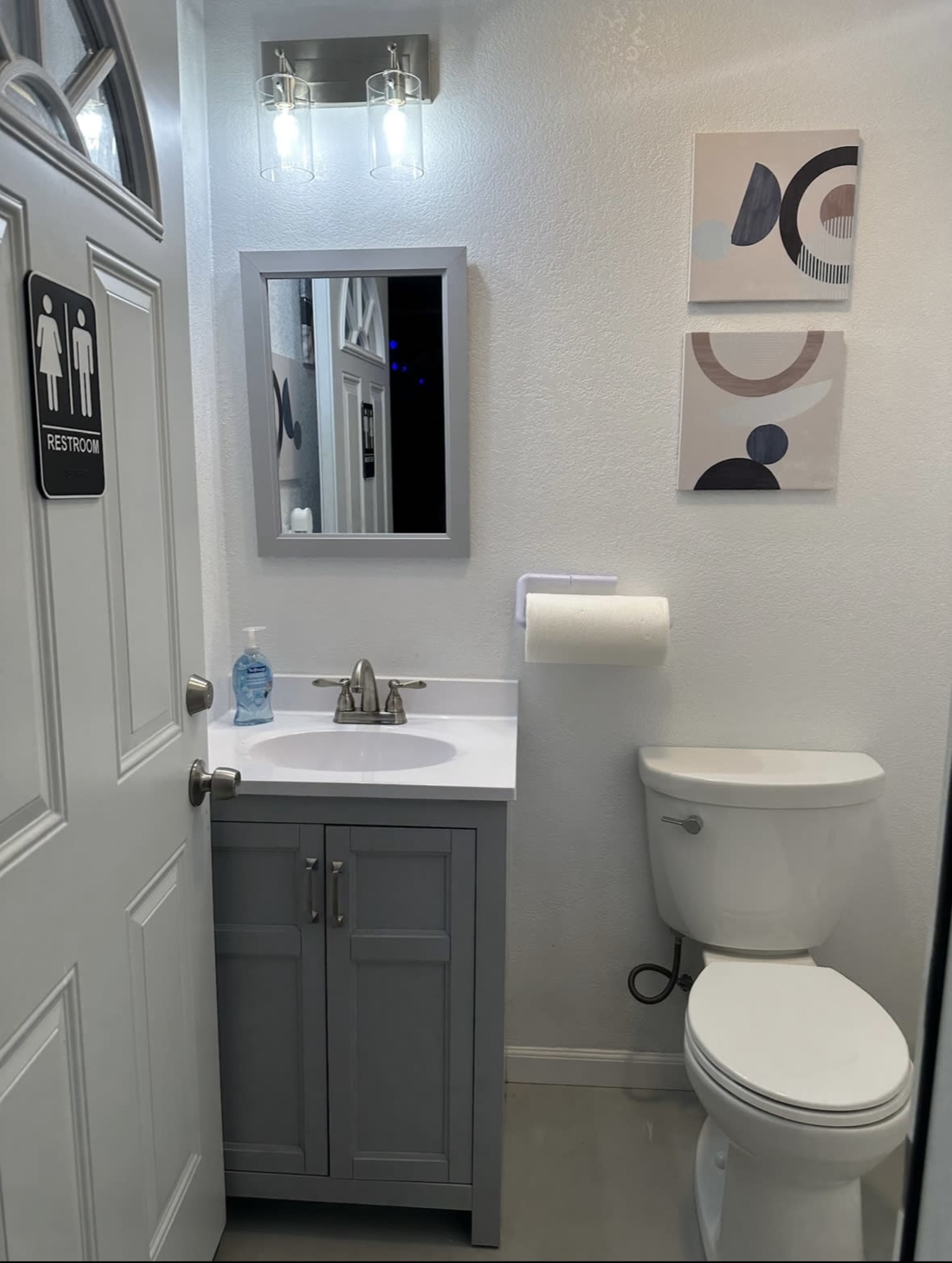 The image shows a small bathroom featuring a sink with a cabinet, a toilet, a mirror, and two framed artworks on the wall.