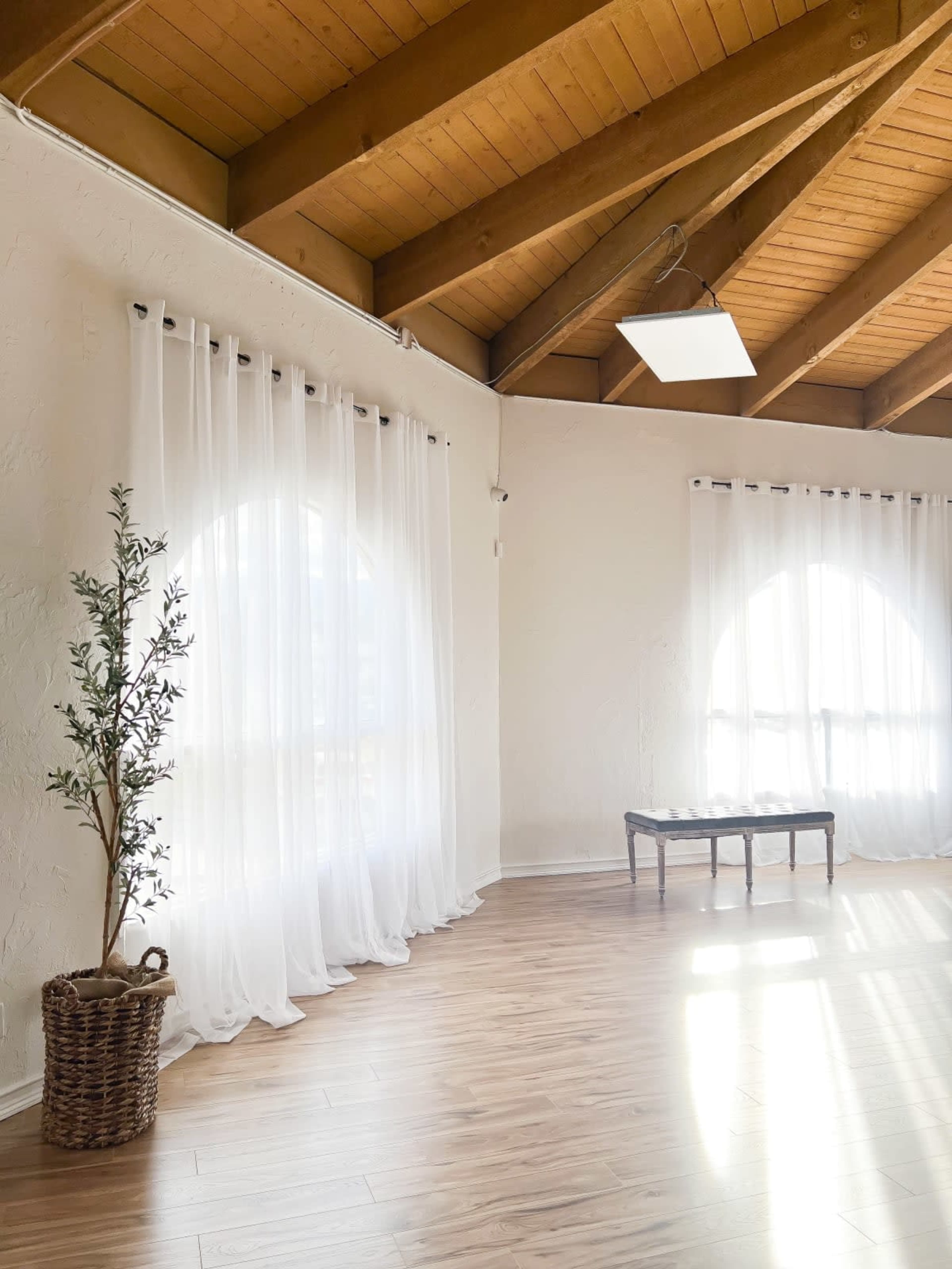 A spacious interior features large windows with sheer curtains, wooden beams, and a simple bench beside a potted plant.