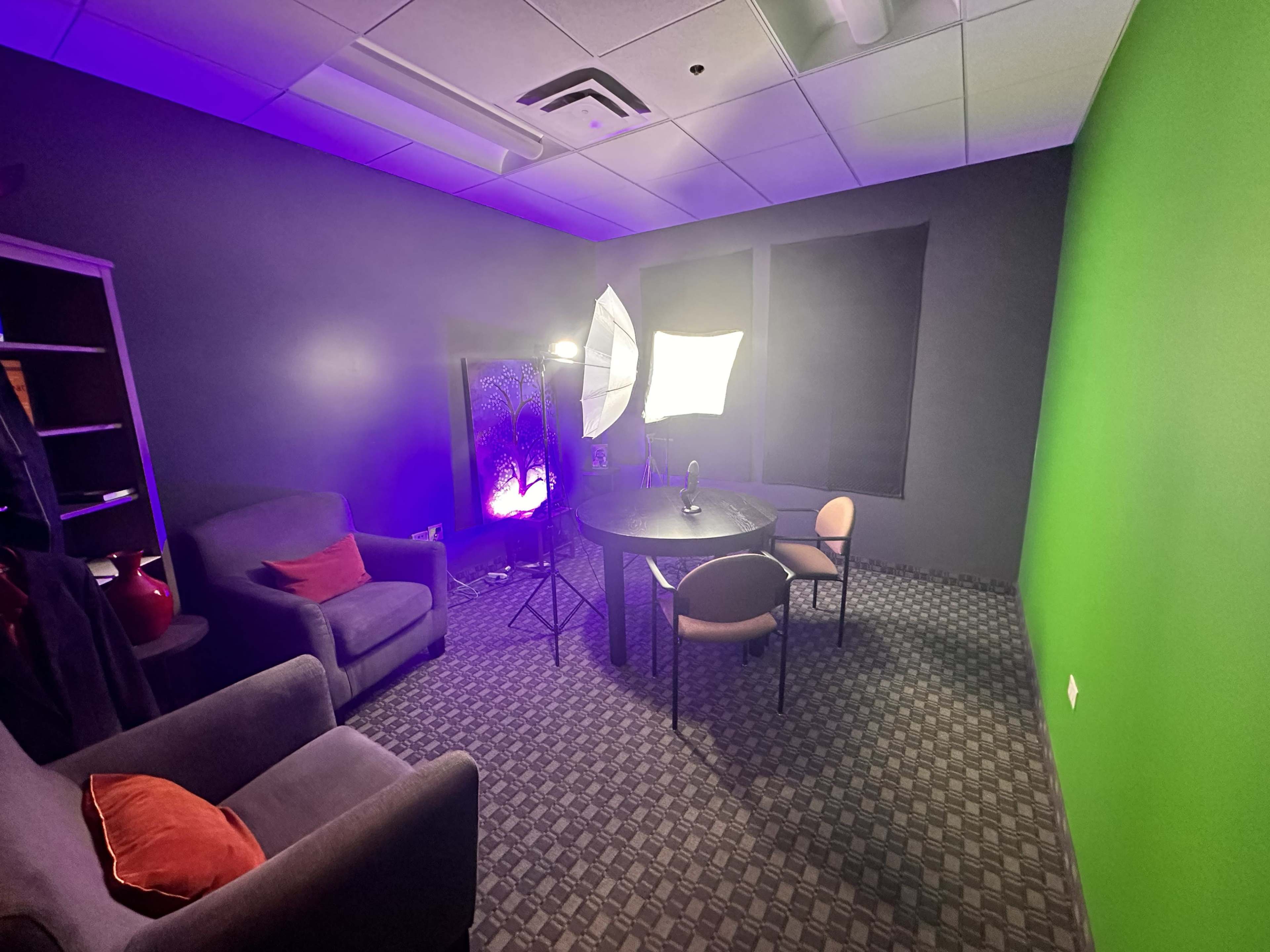 A small studio room with a green screen, a round table, two chairs, and softbox lights positioned for video recording.
