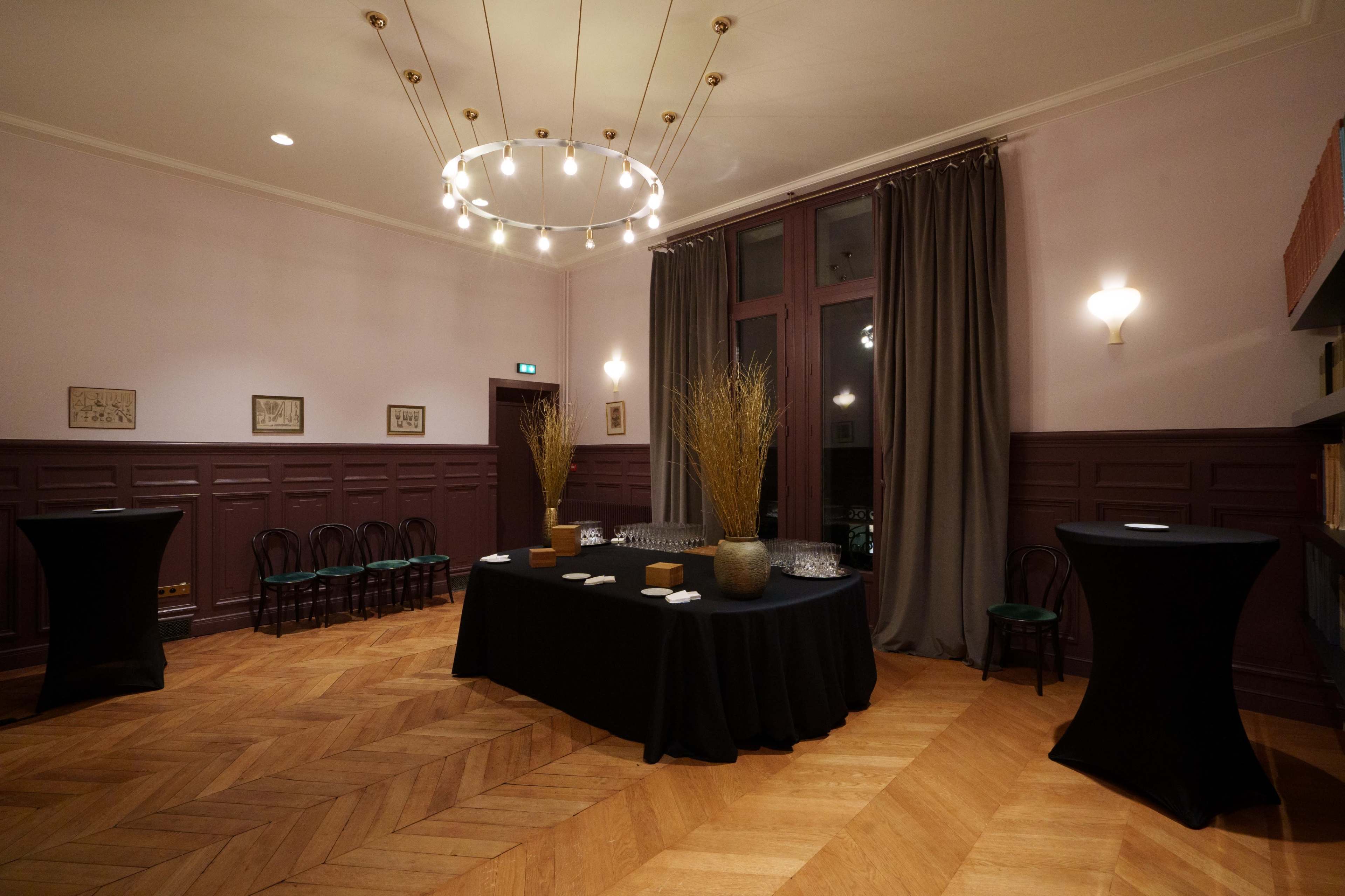 A spacious event room features dark wood flooring, a large black table set with drinks and decorative elements, and tall windows draped with heavy curtains.
