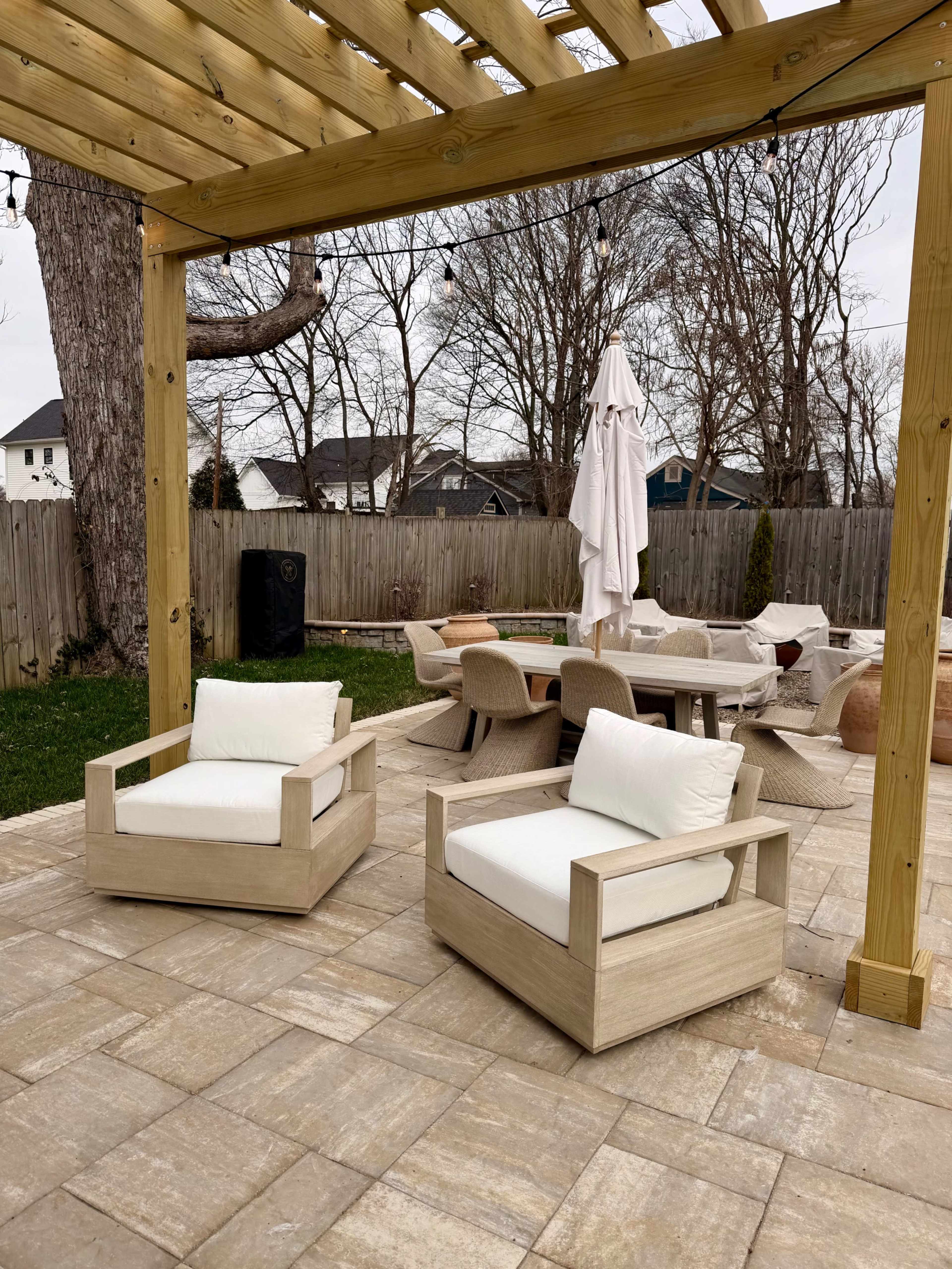 The image shows an outdoor patio area featuring two minimalist wooden armchairs, a dining set with a large umbrella, and a stone paved floor under a wooden pergola.
