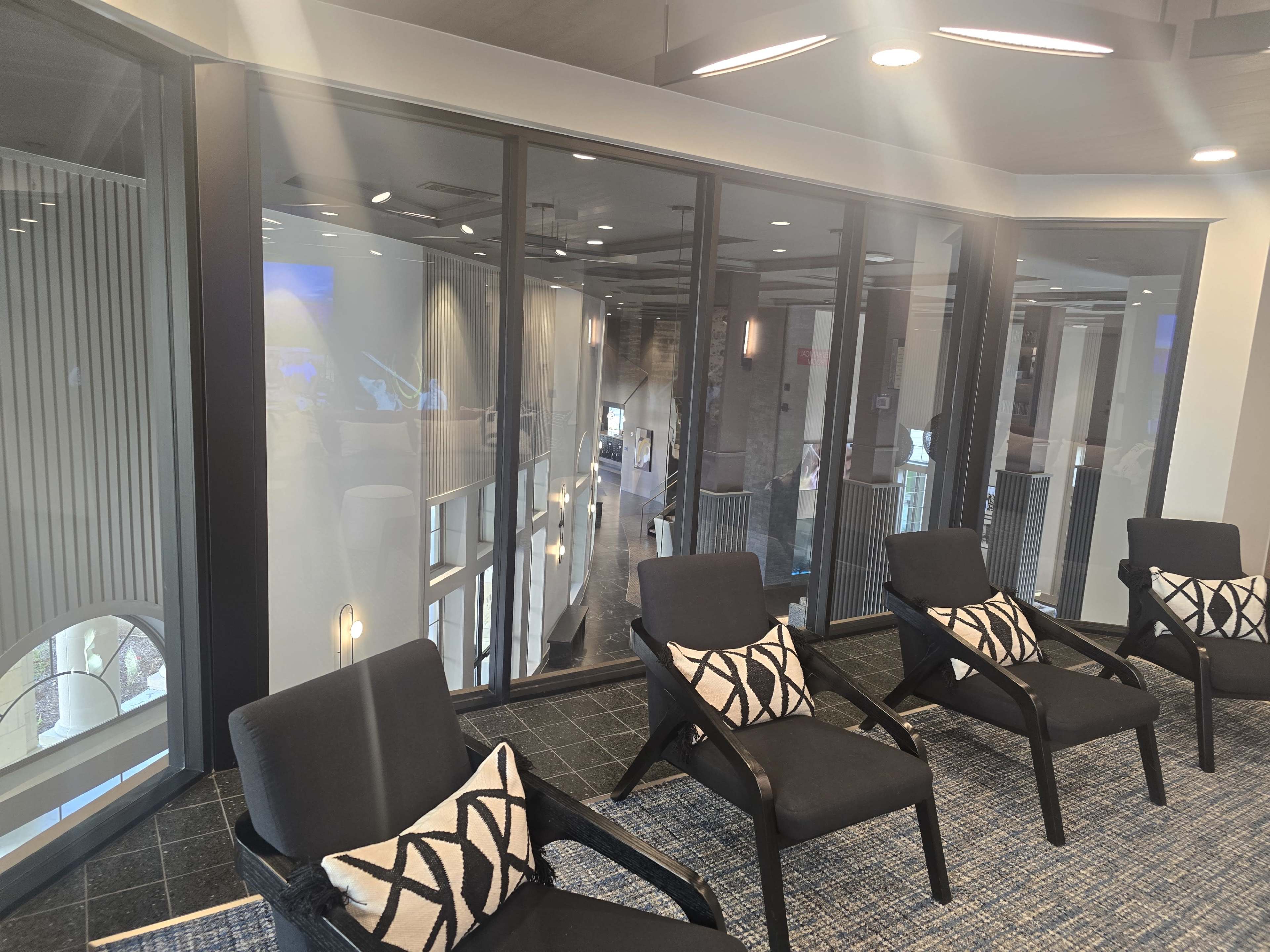 The image shows a modern lounge area with black chairs and large glass windows dividing the space from another room.