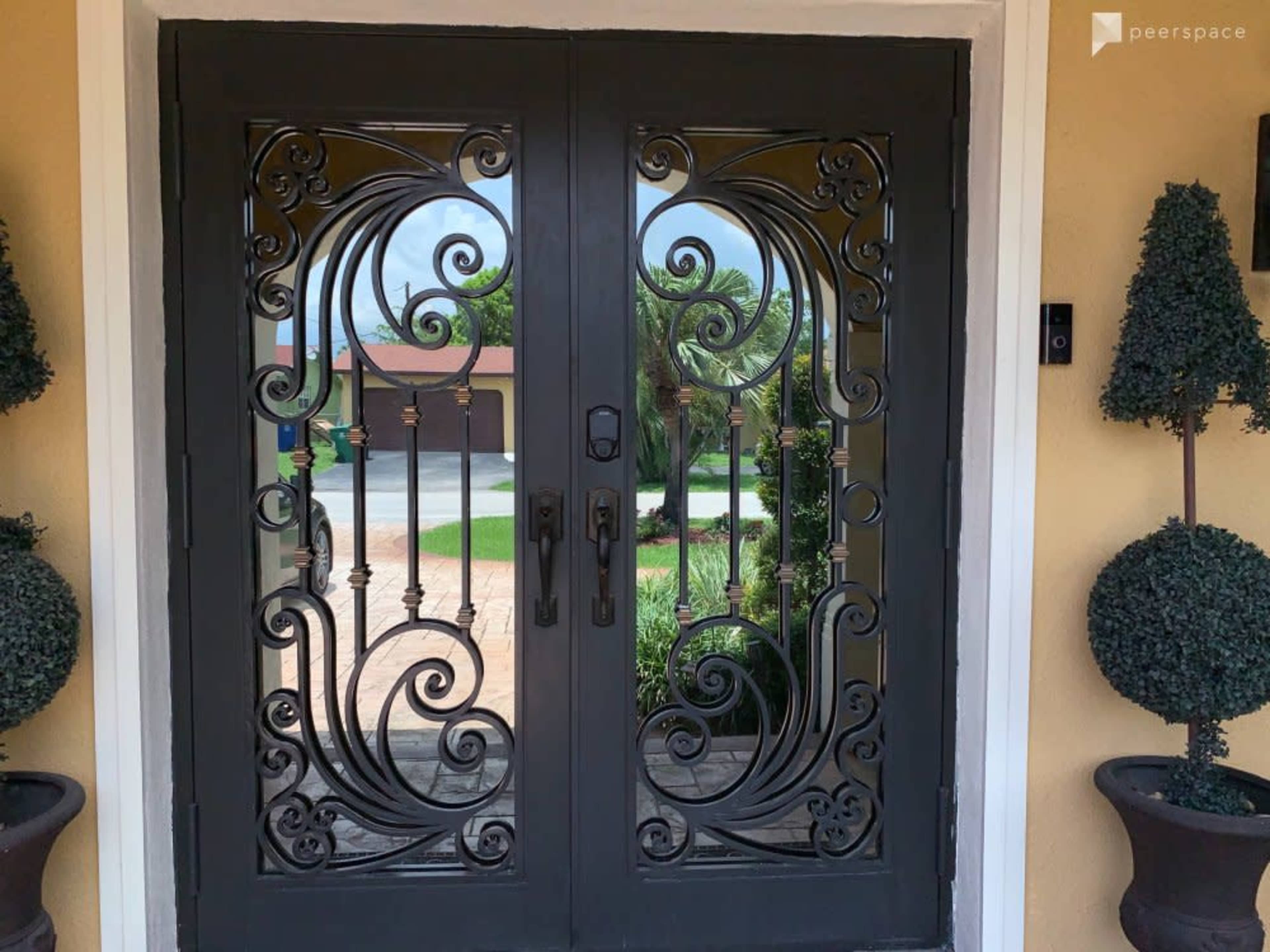 The image shows a pair of ornate double doors with intricate ironwork designs set against a yellow wall, flanked by potted topiary plants.