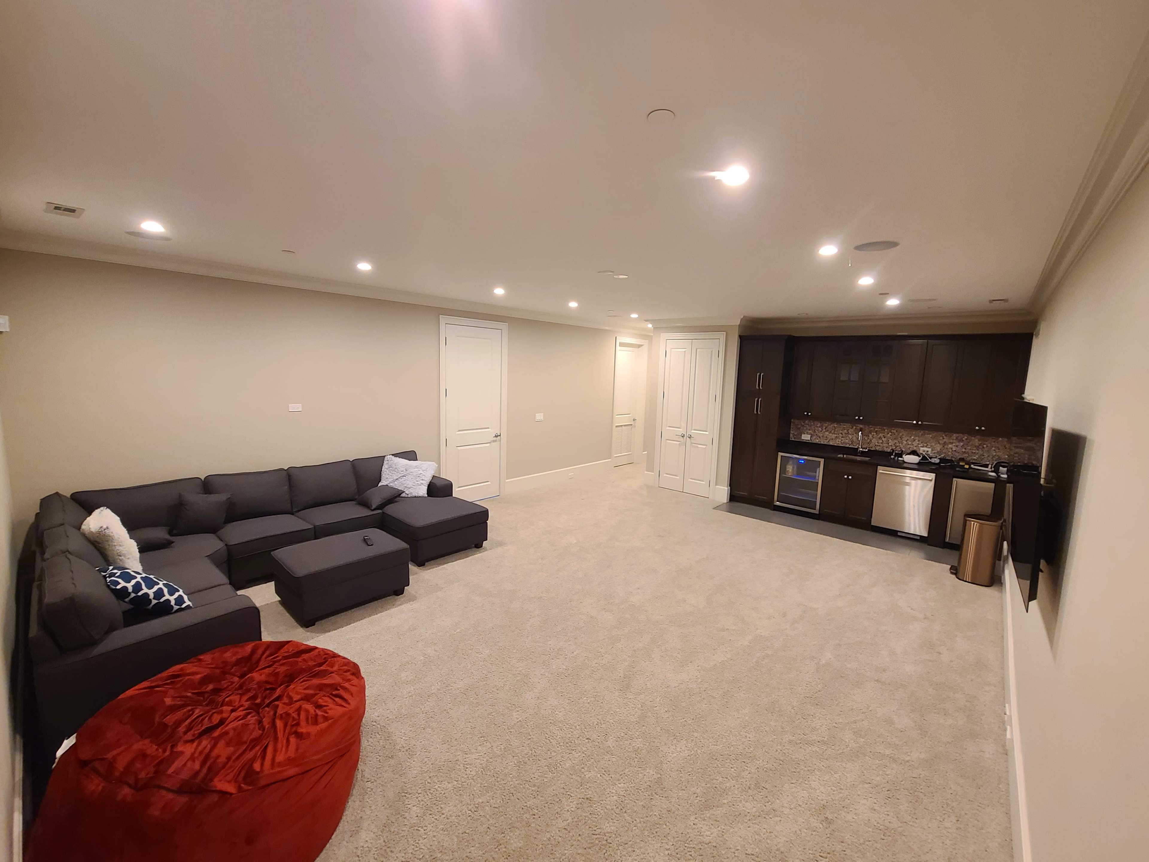 A spacious, carpeted basement with a sectional sofa, a red bean bag chair, and a kitchenette featuring dark cabinetry and appliances.