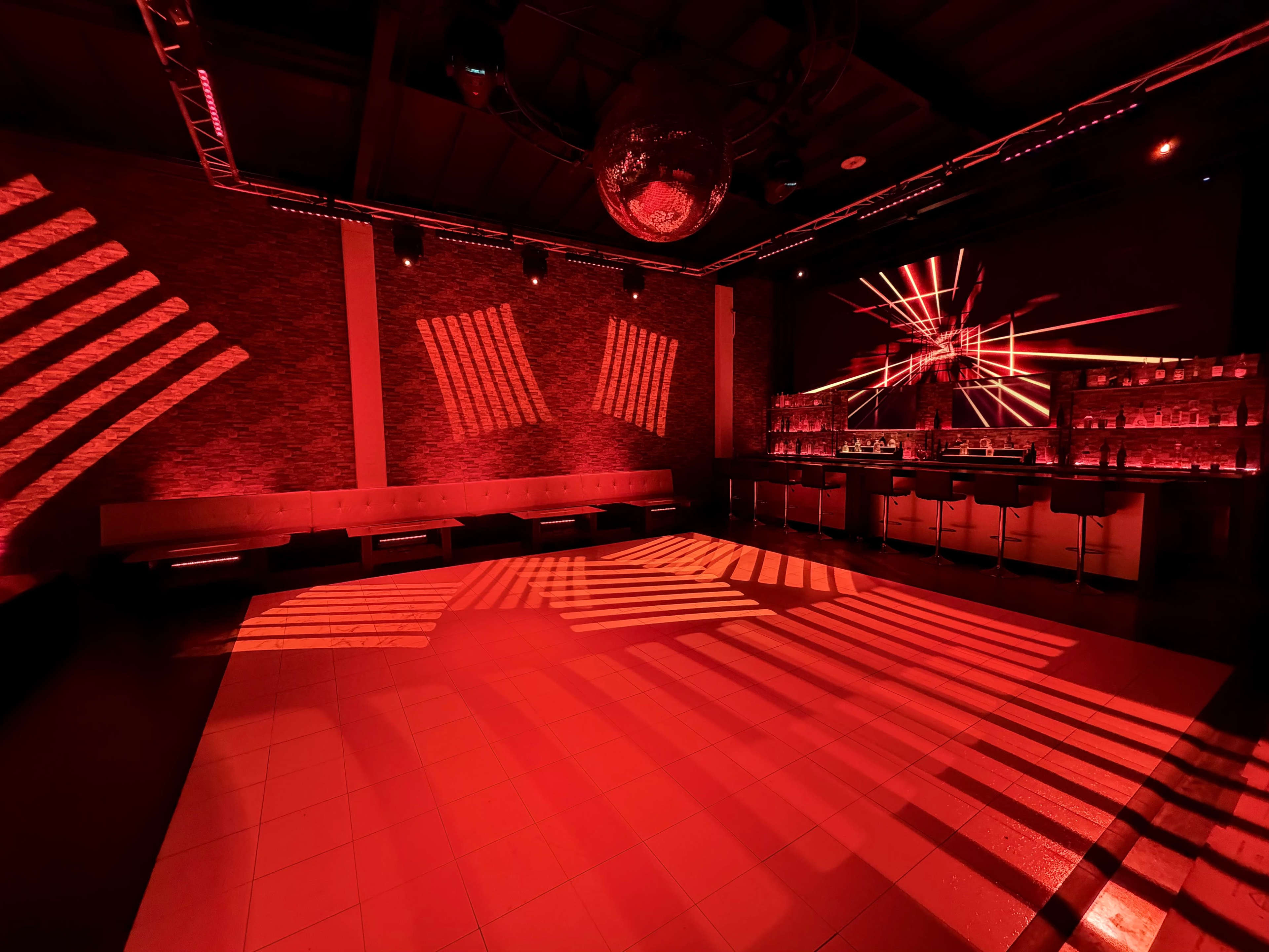 The image shows a dimly lit nightclub interior with red lighting, a bar area, and a disco ball casting patterns on the floor.
