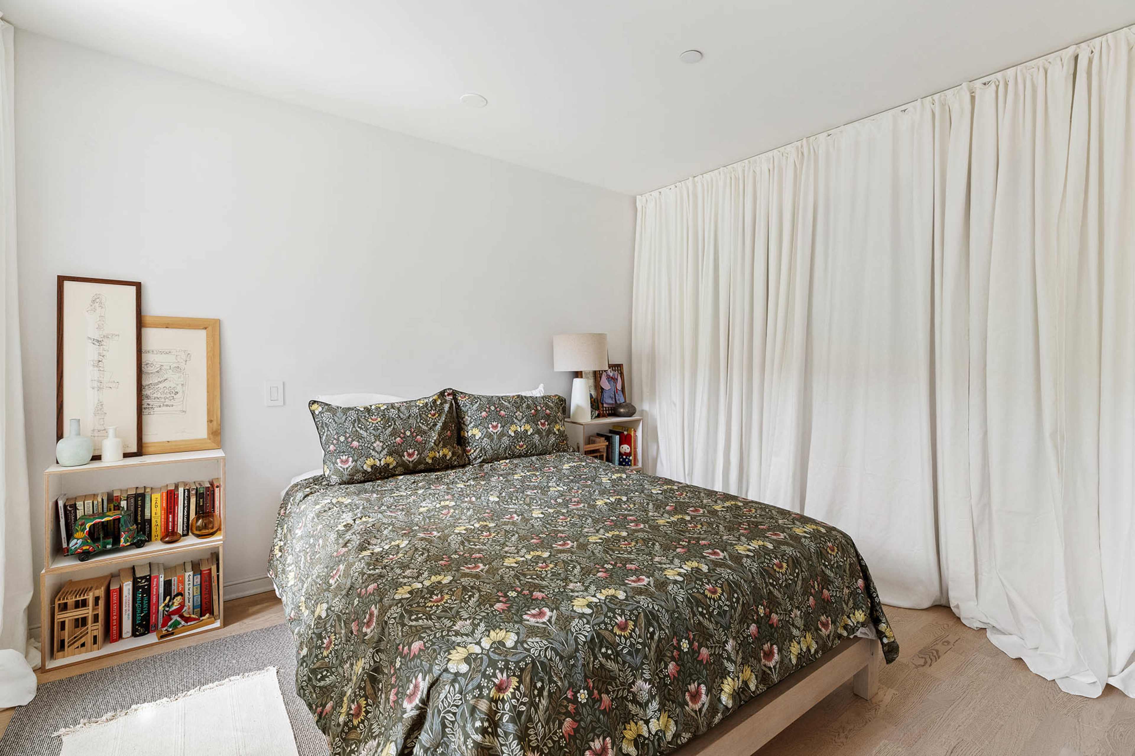 The image shows a bedroom featuring a bed with floral-patterned bedding, a nightstand with a lamp, and a bookshelf filled with books against a wall covered by a white curtain.