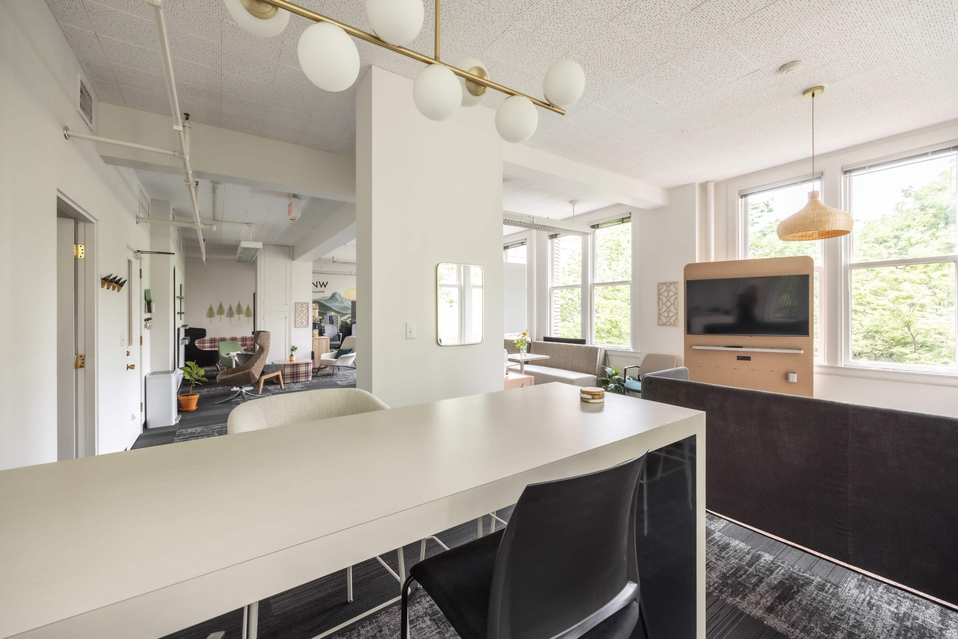 The image shows a modern open-concept office space with a long table, chairs, and large windows allowing natural light.