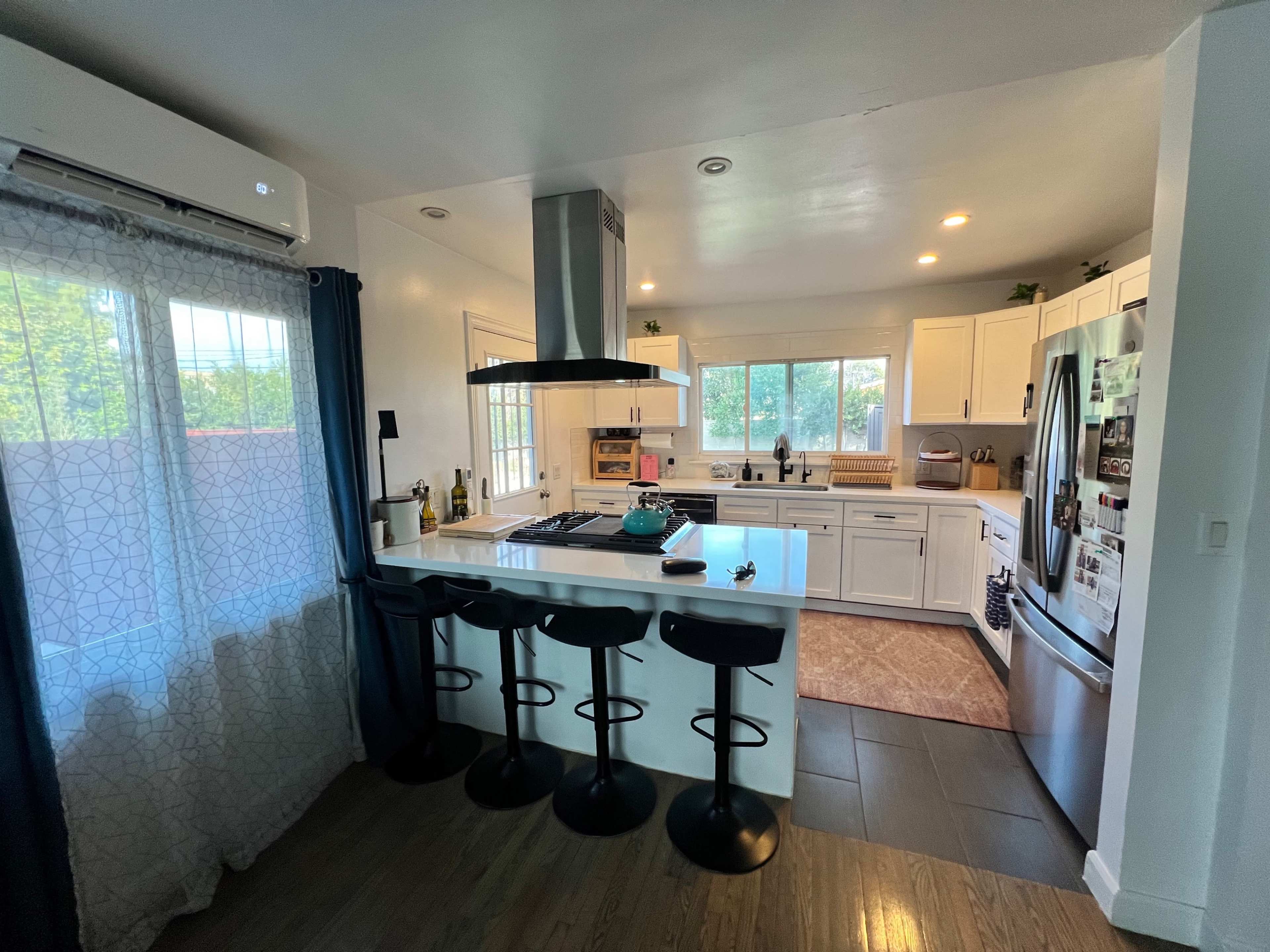 A modern kitchen features white cabinets, a central island with bar stools, and large windows that allow natural light.