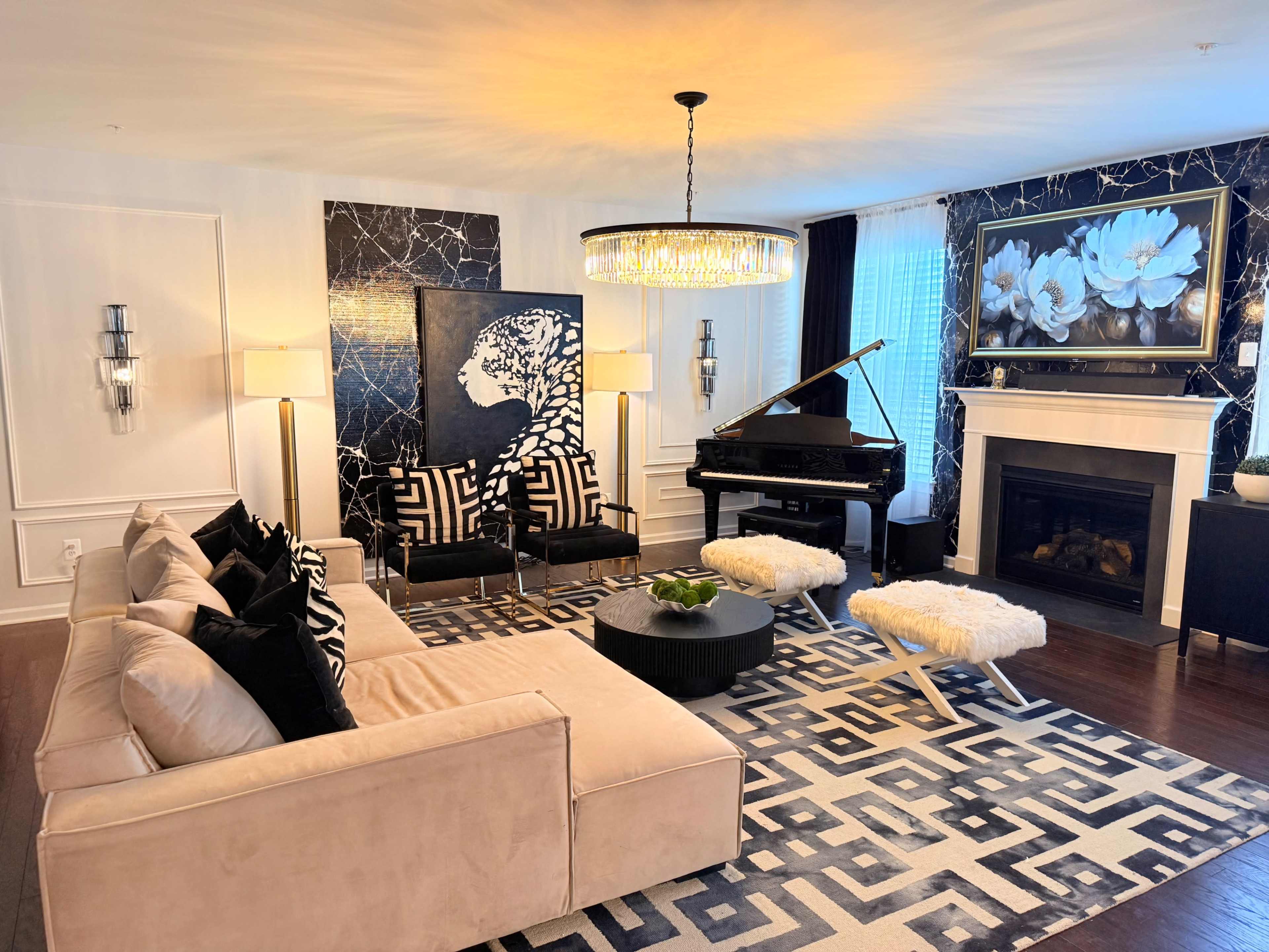 The living room features a beige sofa, black accent chairs, a grand piano, and a patterned rug, all set against a backdrop of a fireplace and decorative wall art.