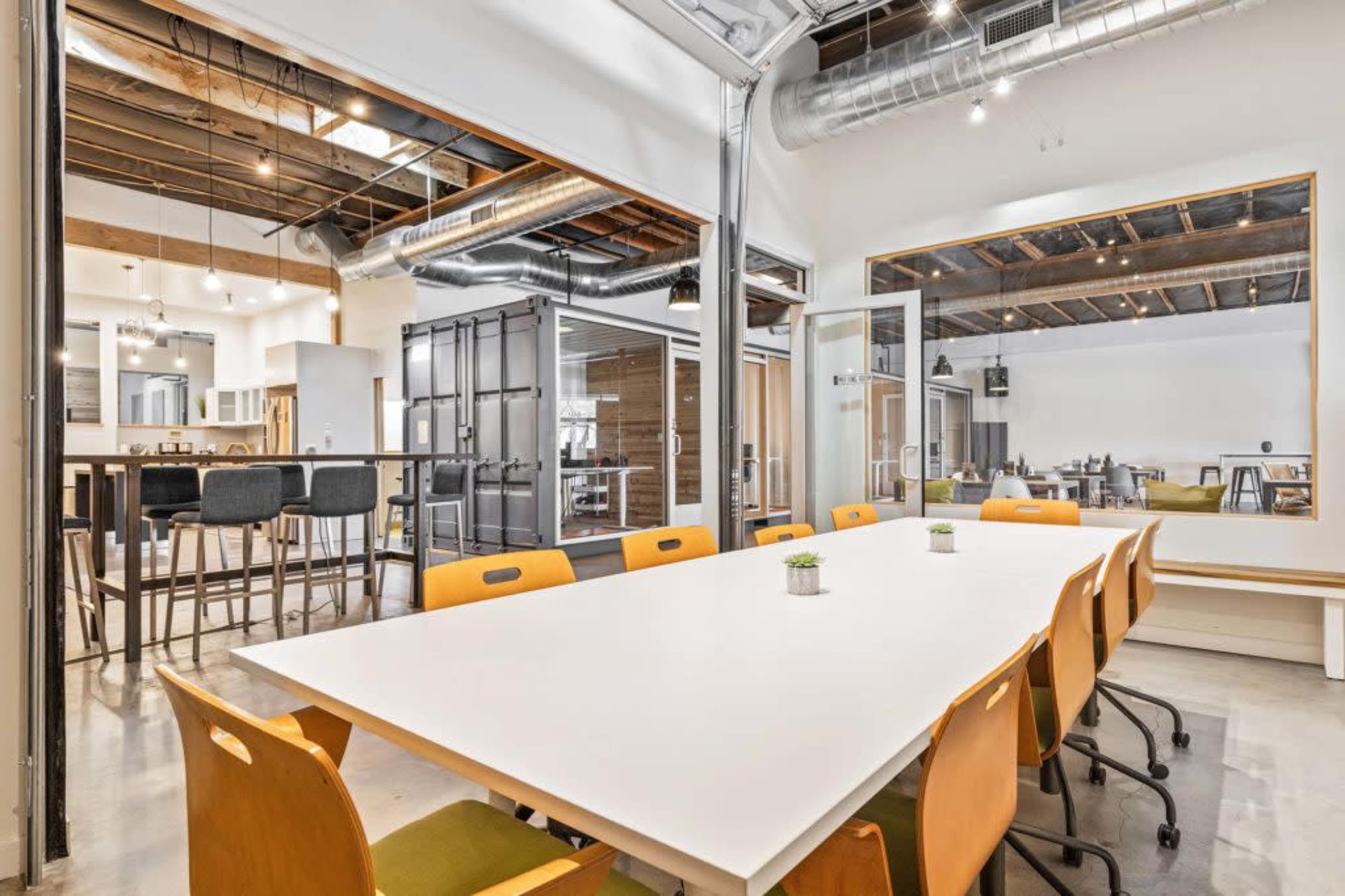A large, well-lit conference room features a white table surrounded by orange chairs and glass-walled offices in the background.