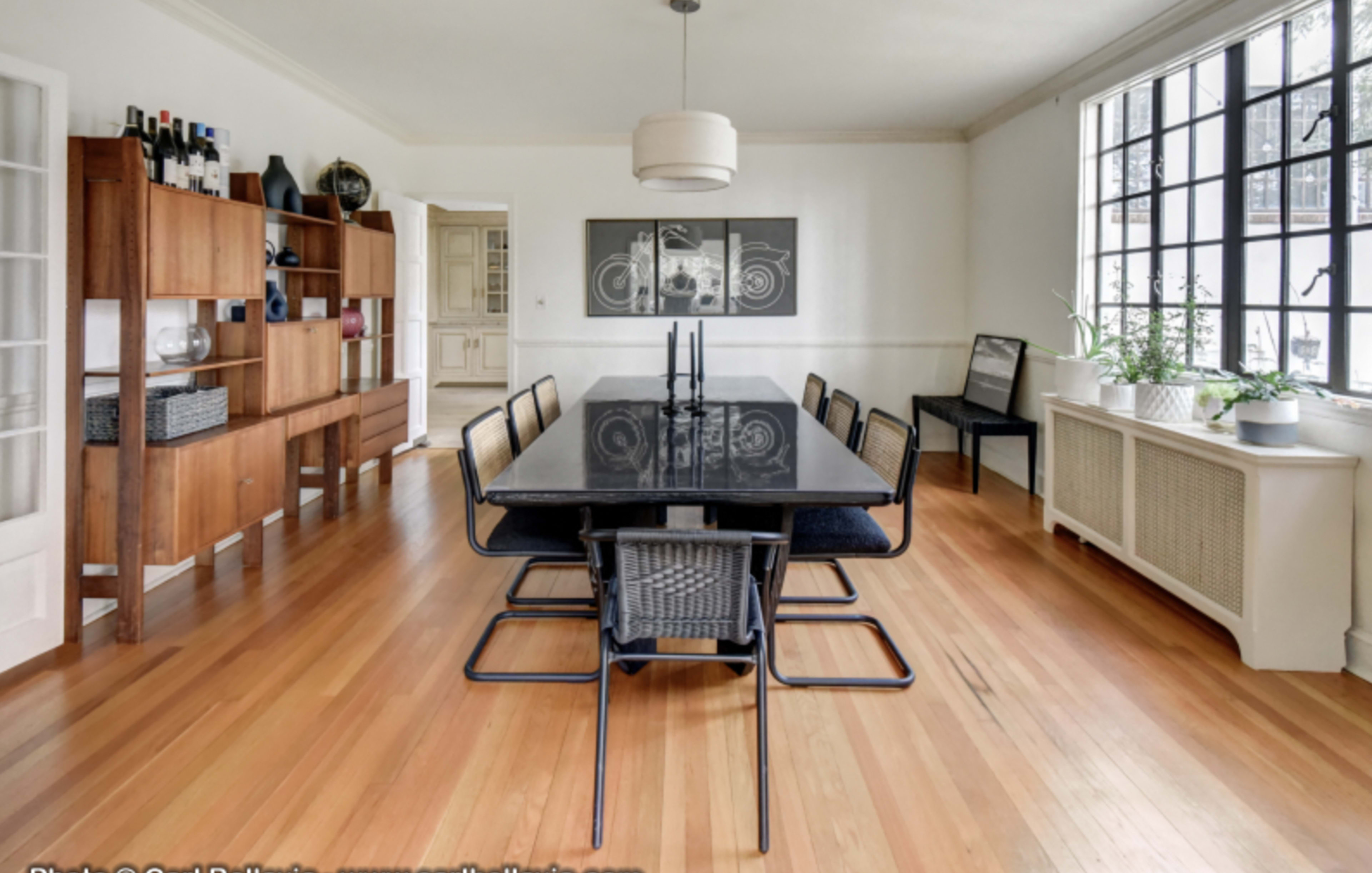 A spacious dining room features a long black table with metal and wicker chairs, hardwood floors, and a large window allowing natural light to enter.